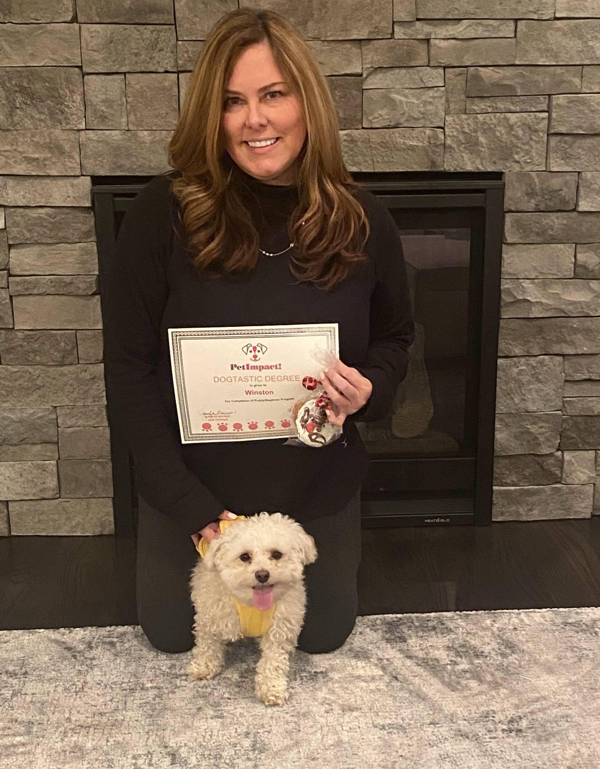 Woman kneeling with small dog; holding certificate in front of fireplace.