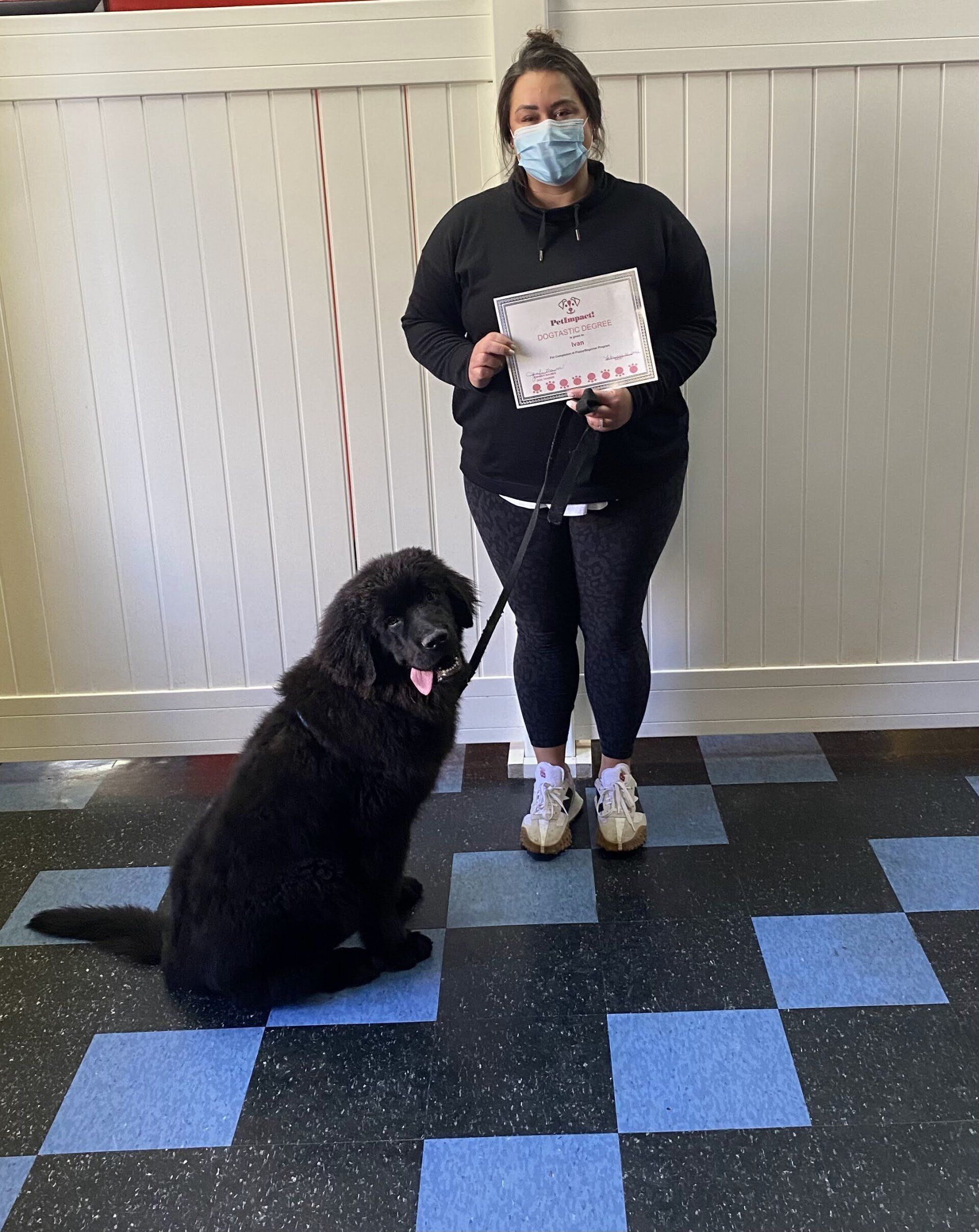 Woman holding a certificate poses with a large black dog, both indoors, against a white wall and checkered floor.
