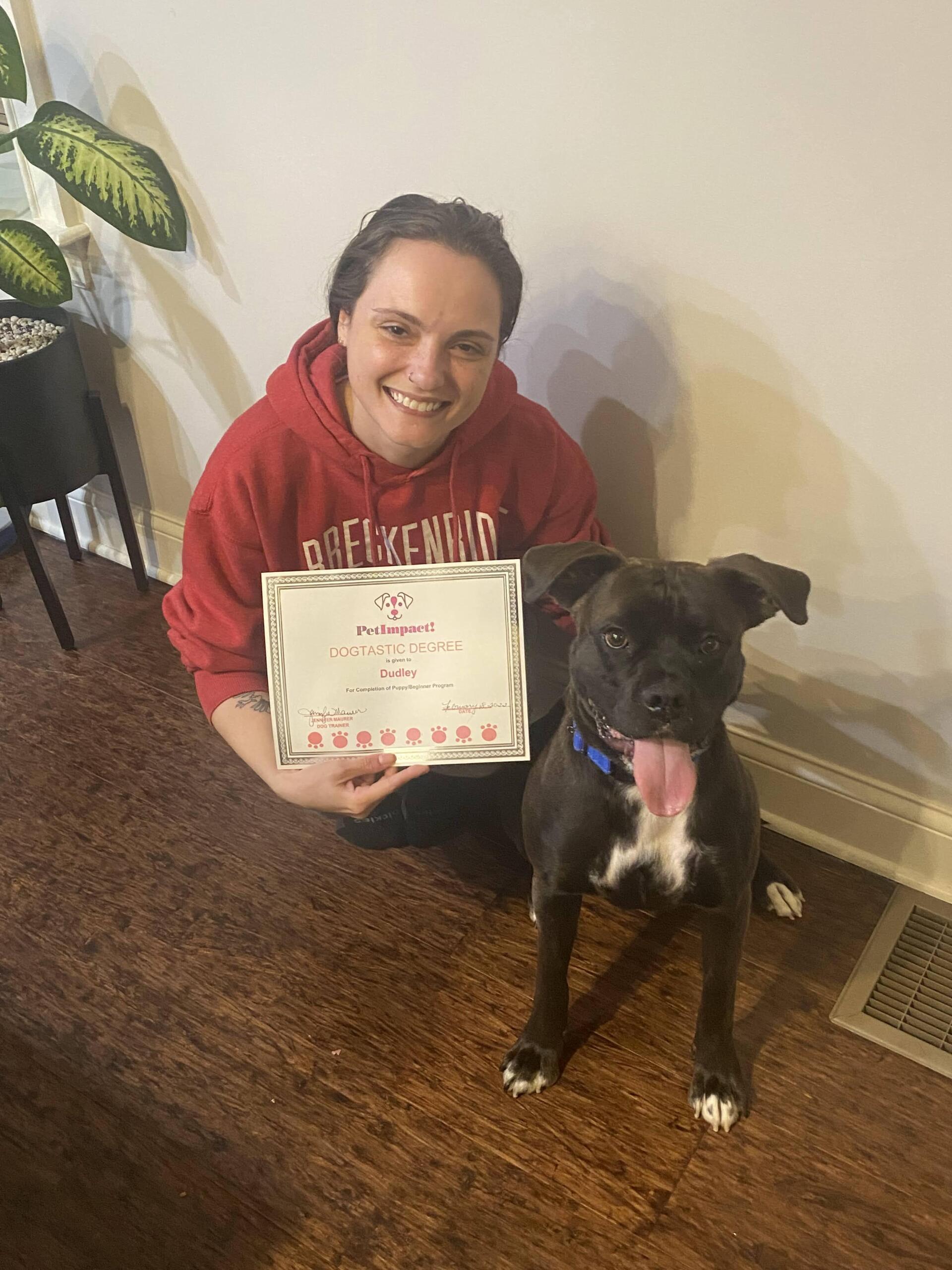 Woman in red hoodie kneels with a dog, both smiling, holding a certificate. Brown floor, white wall.