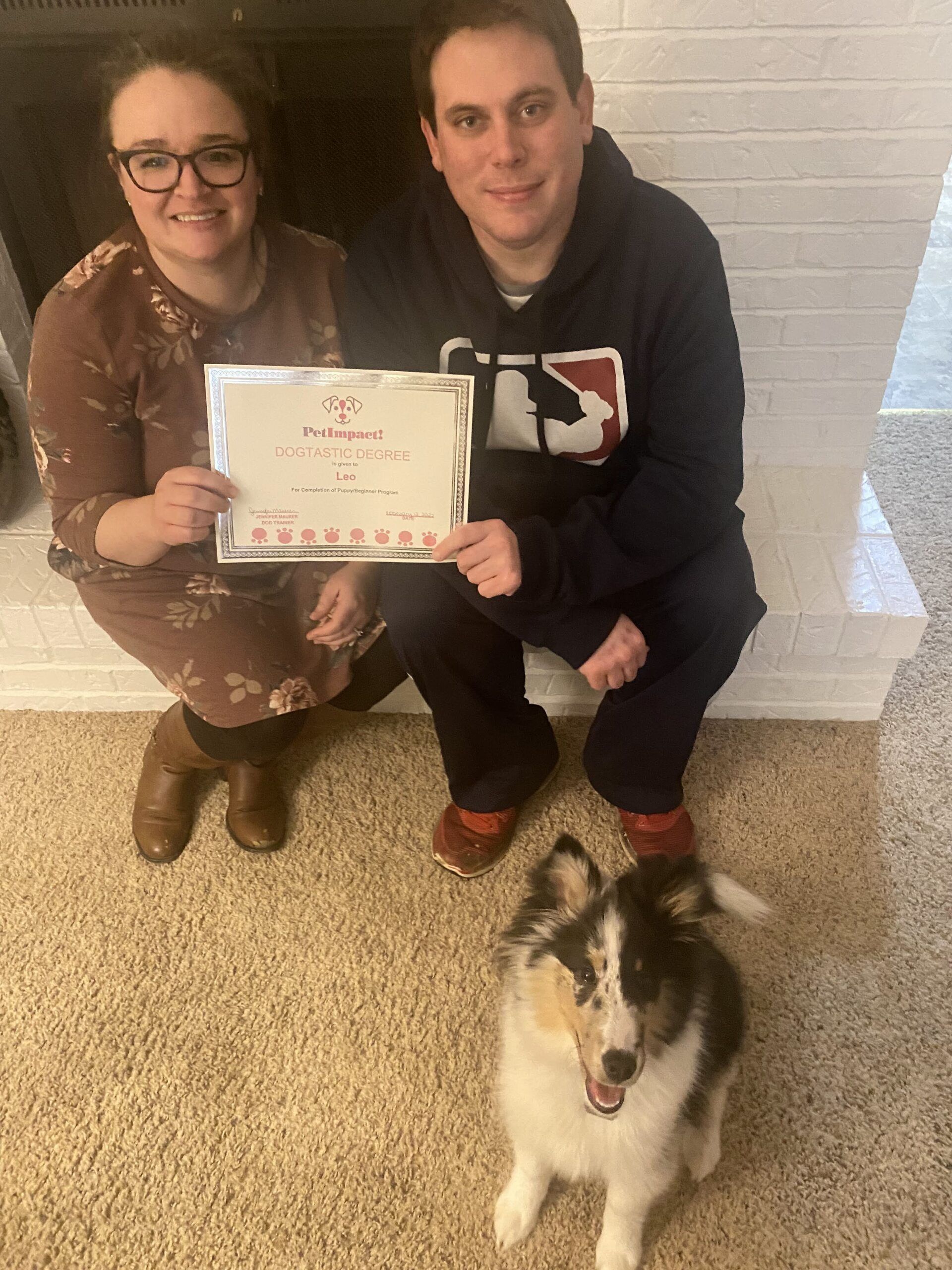 Couple and dog with certificate, kneeling near fireplace. Woman smiling and wearing glasses; man in sweatshirt.
