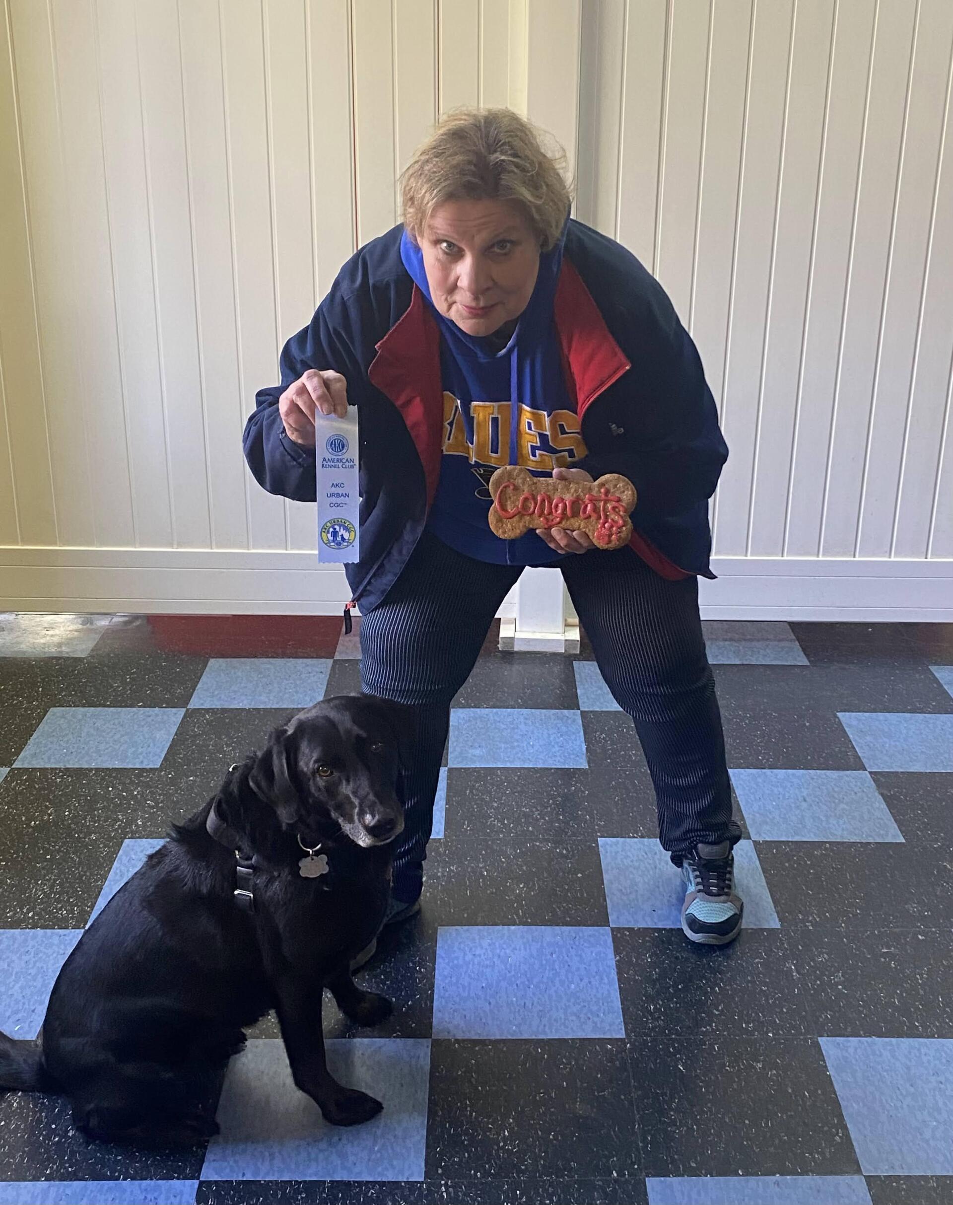Person with dog, holding ribbon and medal. Person in blue jacket, dog is black, sitting on checkered floor.