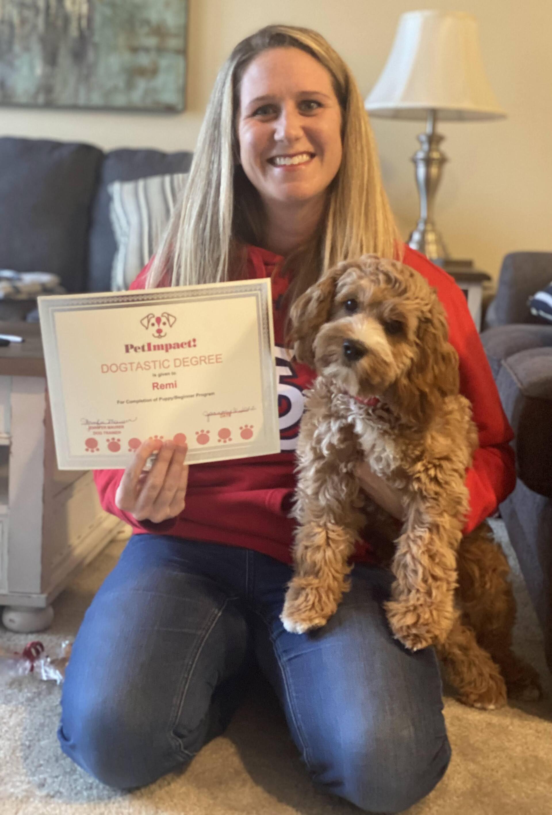 Woman kneels, smiles, holding a puppy and certificate in a living room.