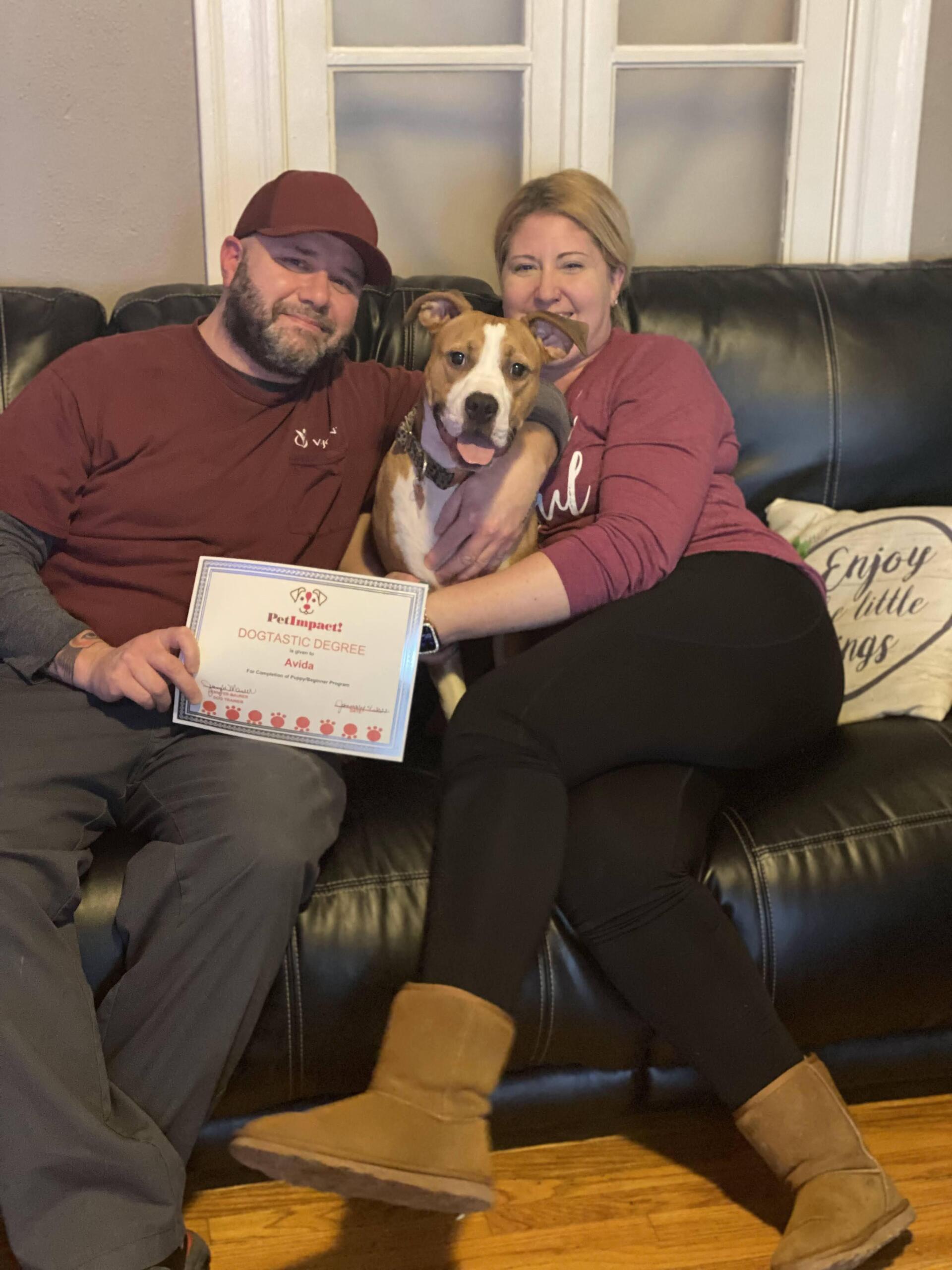 Couple and dog sit on couch holding certificate. Brown and white dog in middle.