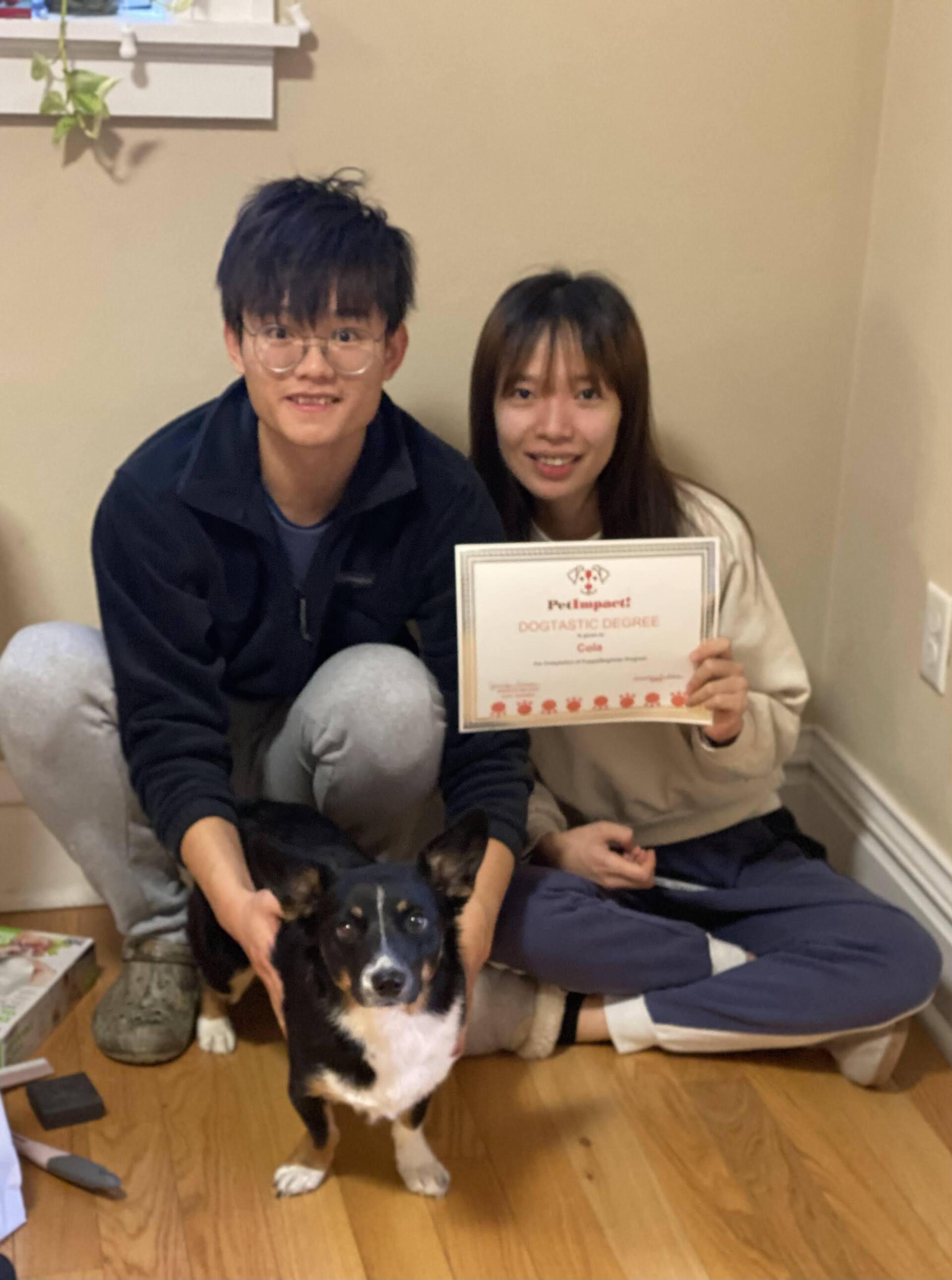 Two people and a dog seated on the floor, holding a certificate. They are smiling. Indoor setting.