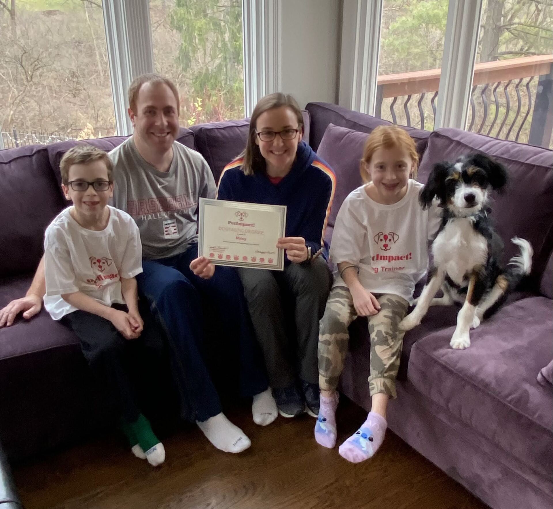Family and dog sit on a purple couch, holding a certificate; natural light through windows.