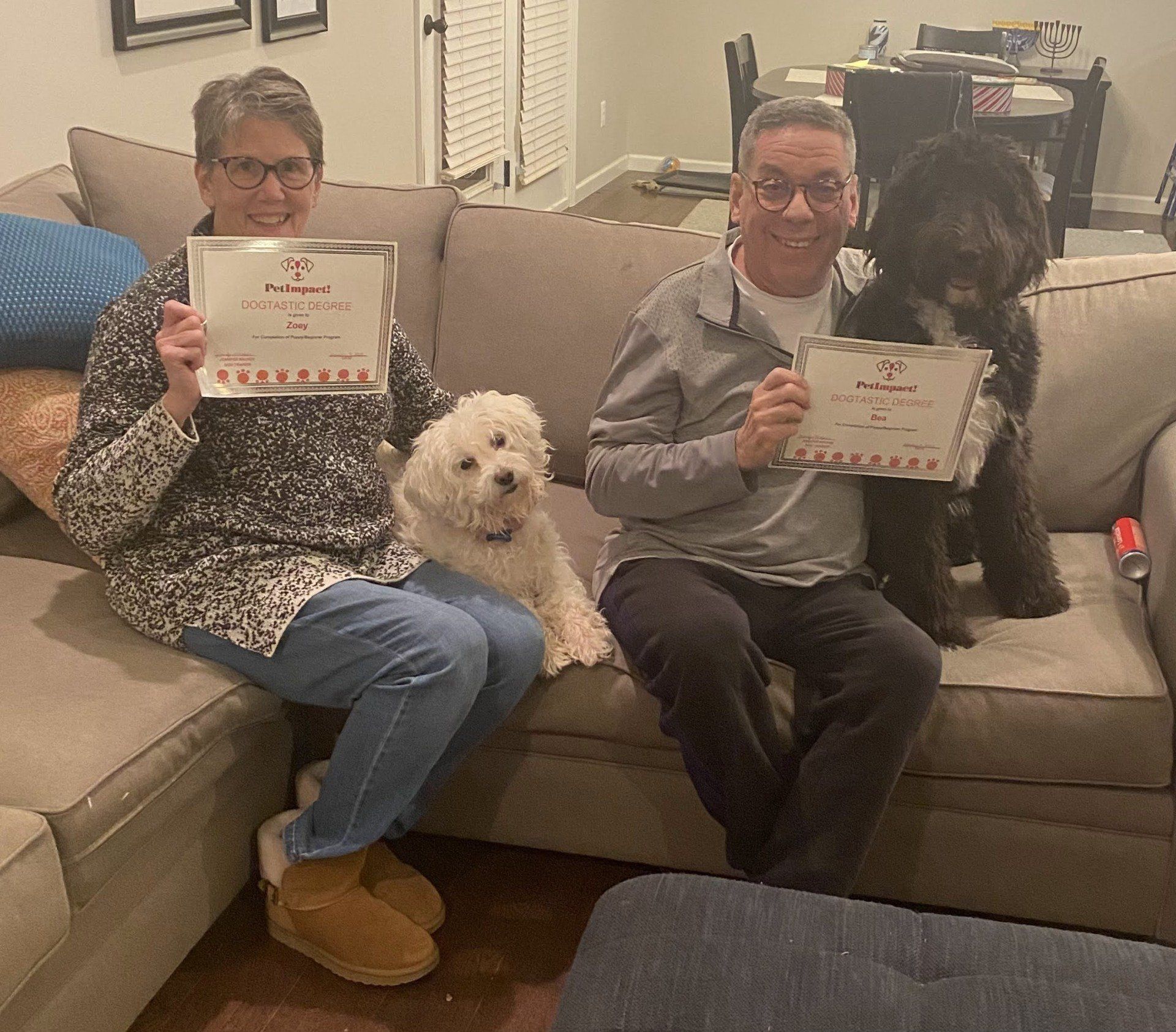 Two people on a couch with two dogs, holding certificates. Happy expressions. Living room setting.