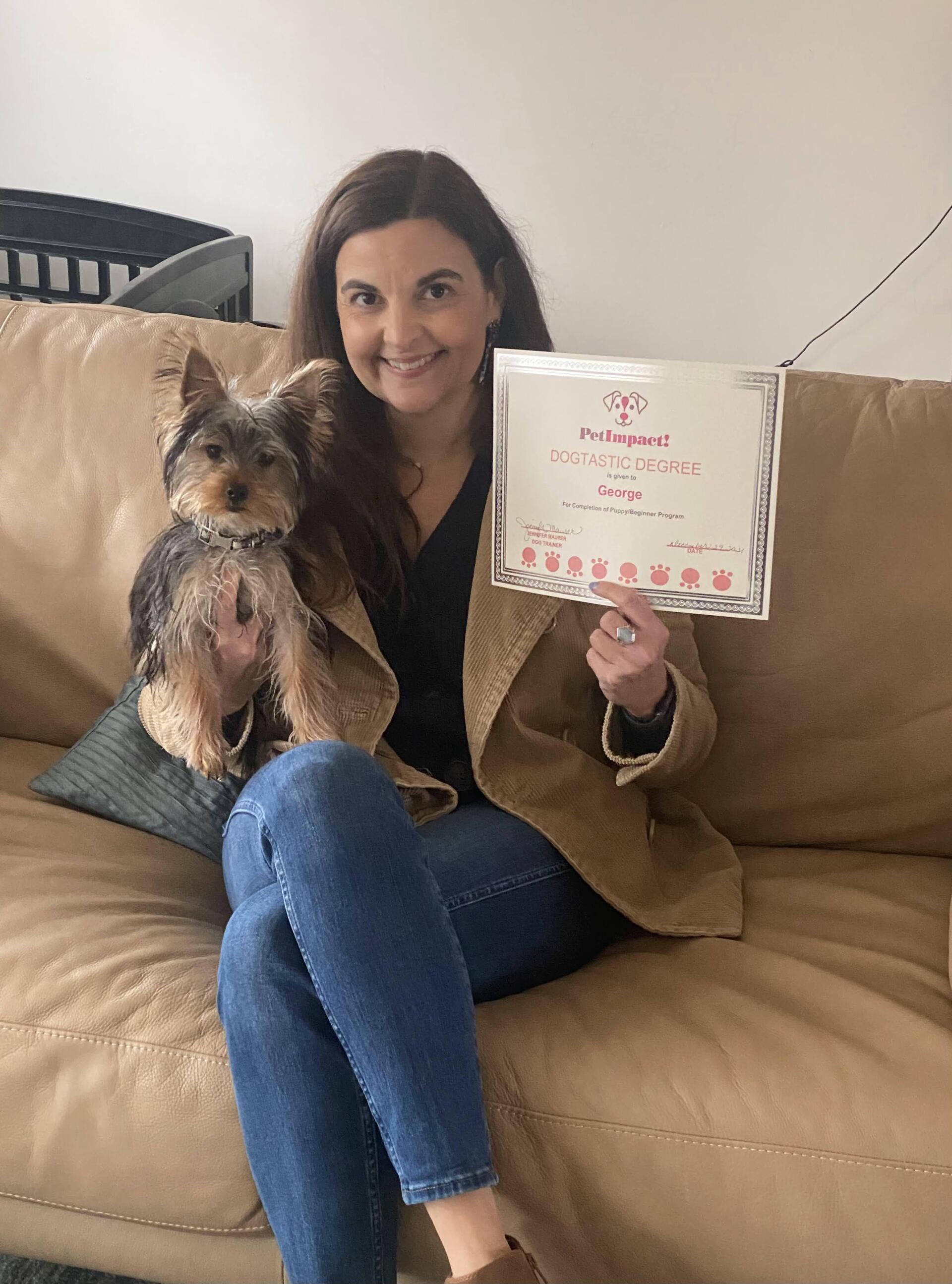 Woman on a couch holding a dog and a certificate; smiling.