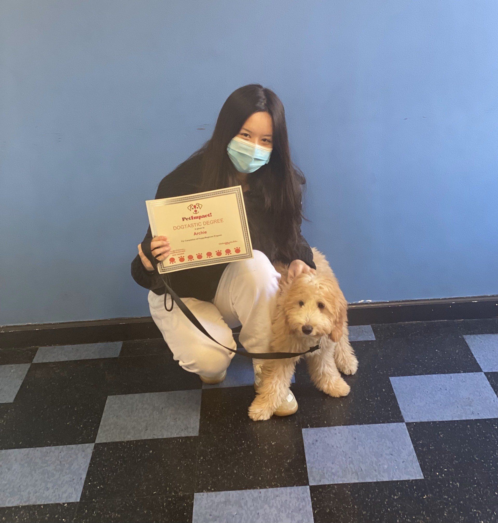 Woman in mask kneels with dog, holding framed certificate. Against a blue wall, black and blue tiled floor.
