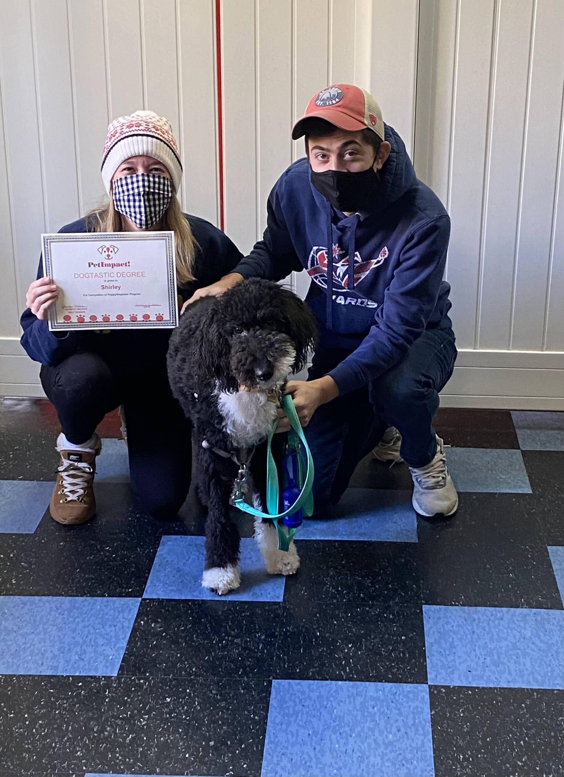 Two people kneel with a fluffy black and white dog holding a certificate.
