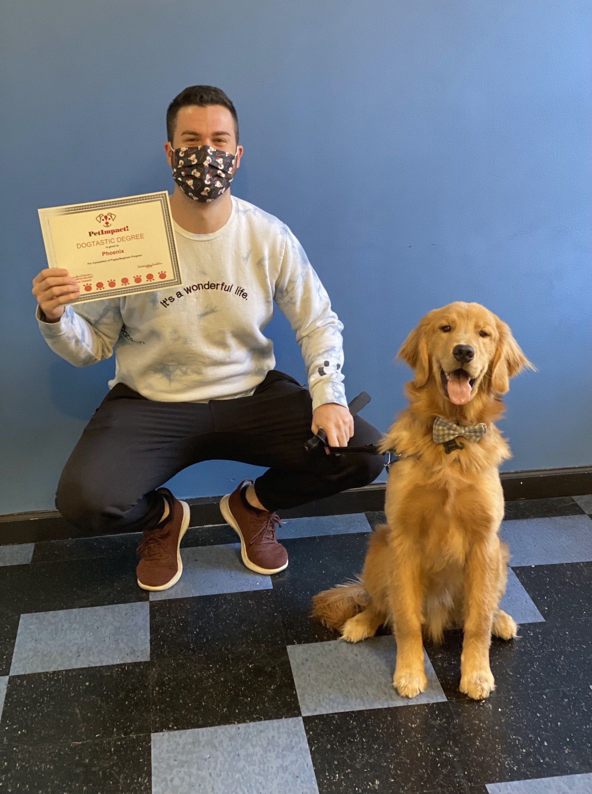 Man in mask squats next to Golden Retriever; both looking at camera. Man holds certificate; setting has blue wall, checkered floor.
