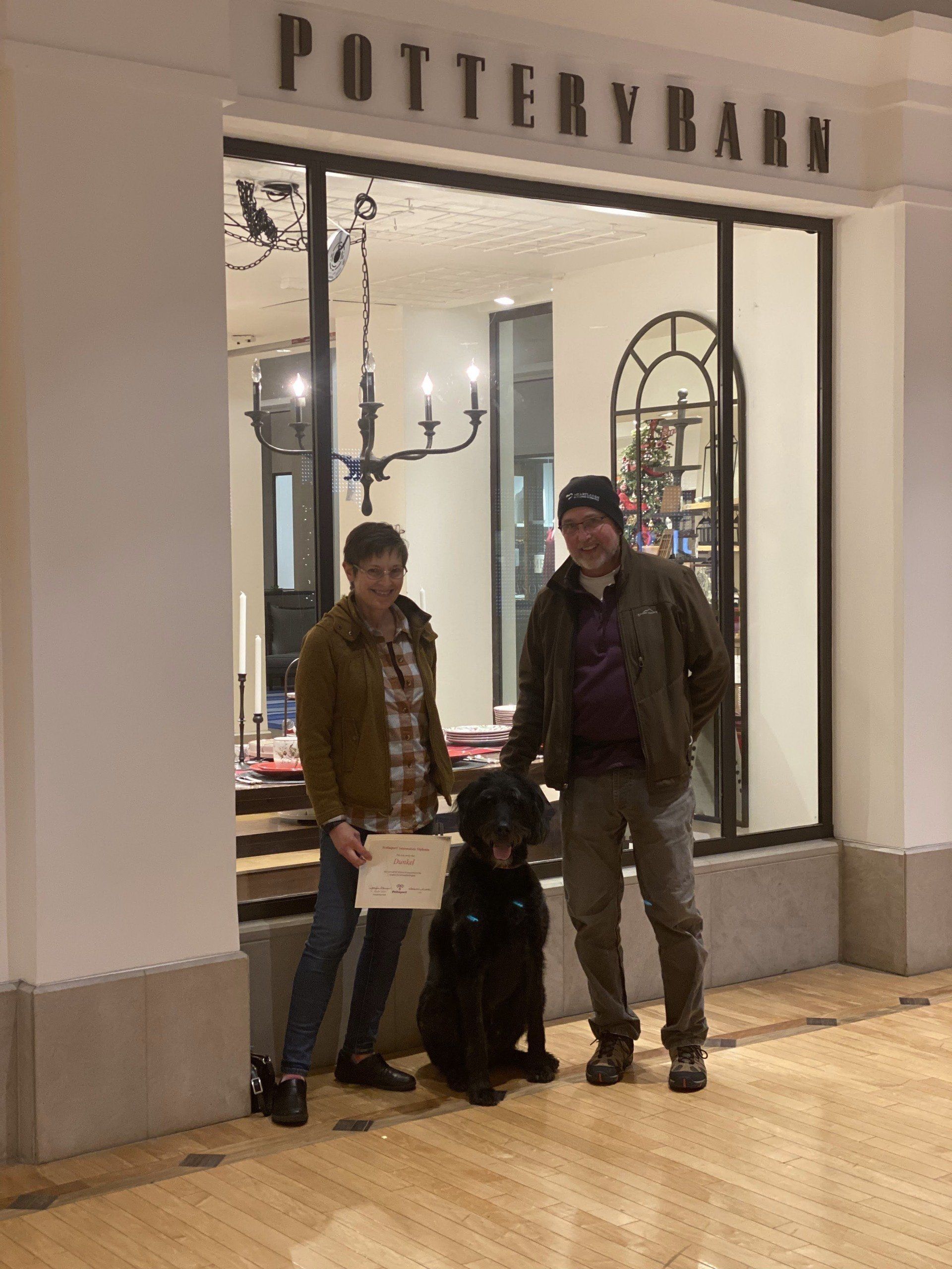 Two people and a black dog pose in front of a Pottery Barn store.