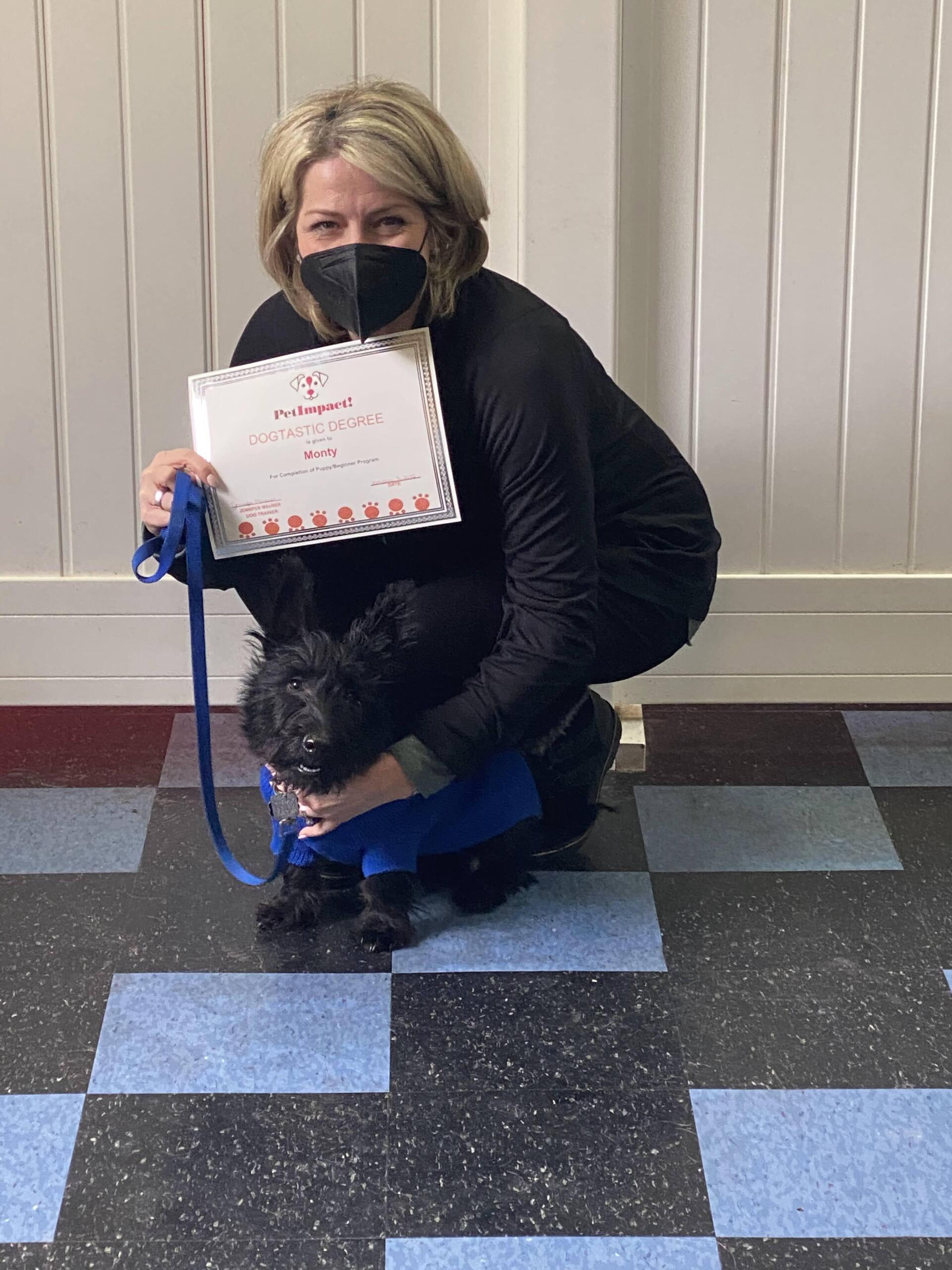 Woman in mask with black dog in blue coat, holding certificate. Sitting on blue and black tile floor.