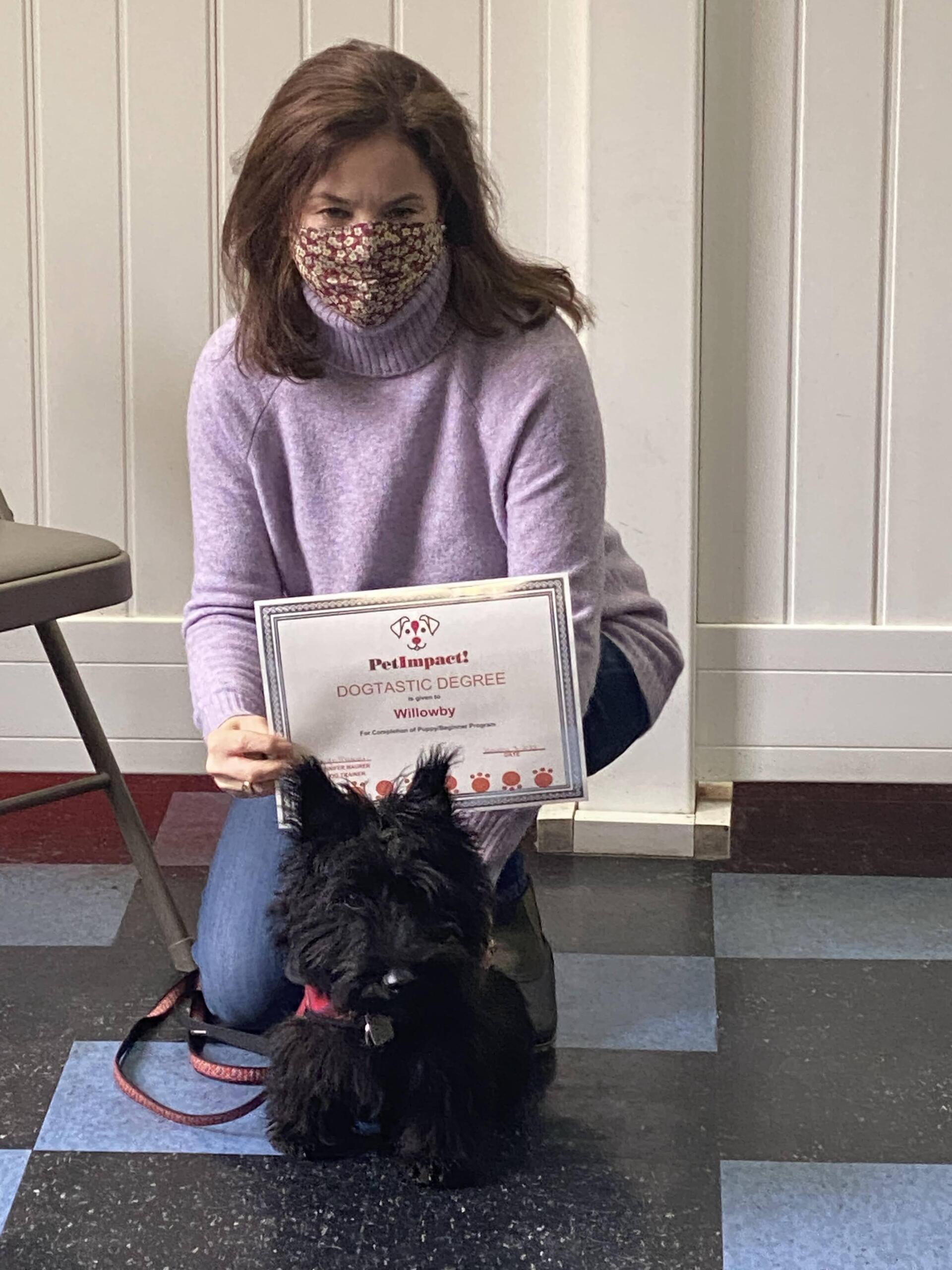 Woman in mask kneeling with black dog, holding certificate.