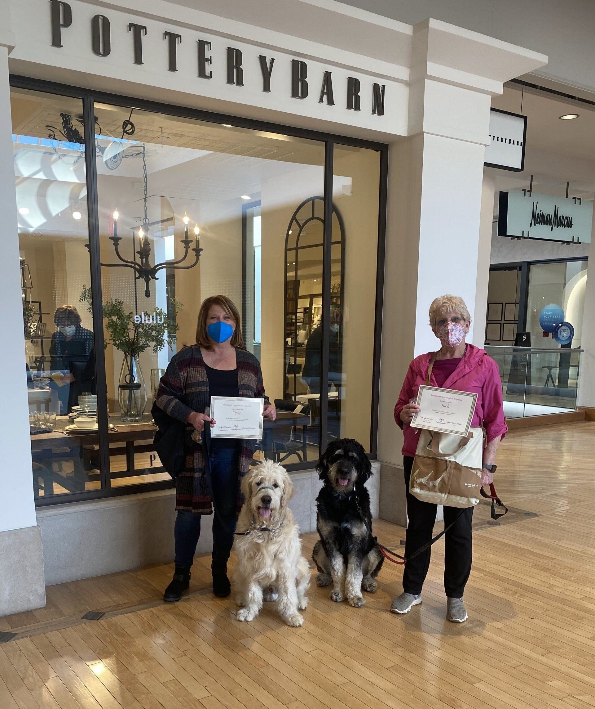 Two women with two dogs pose in front of a Pottery Barn store, holding papers.