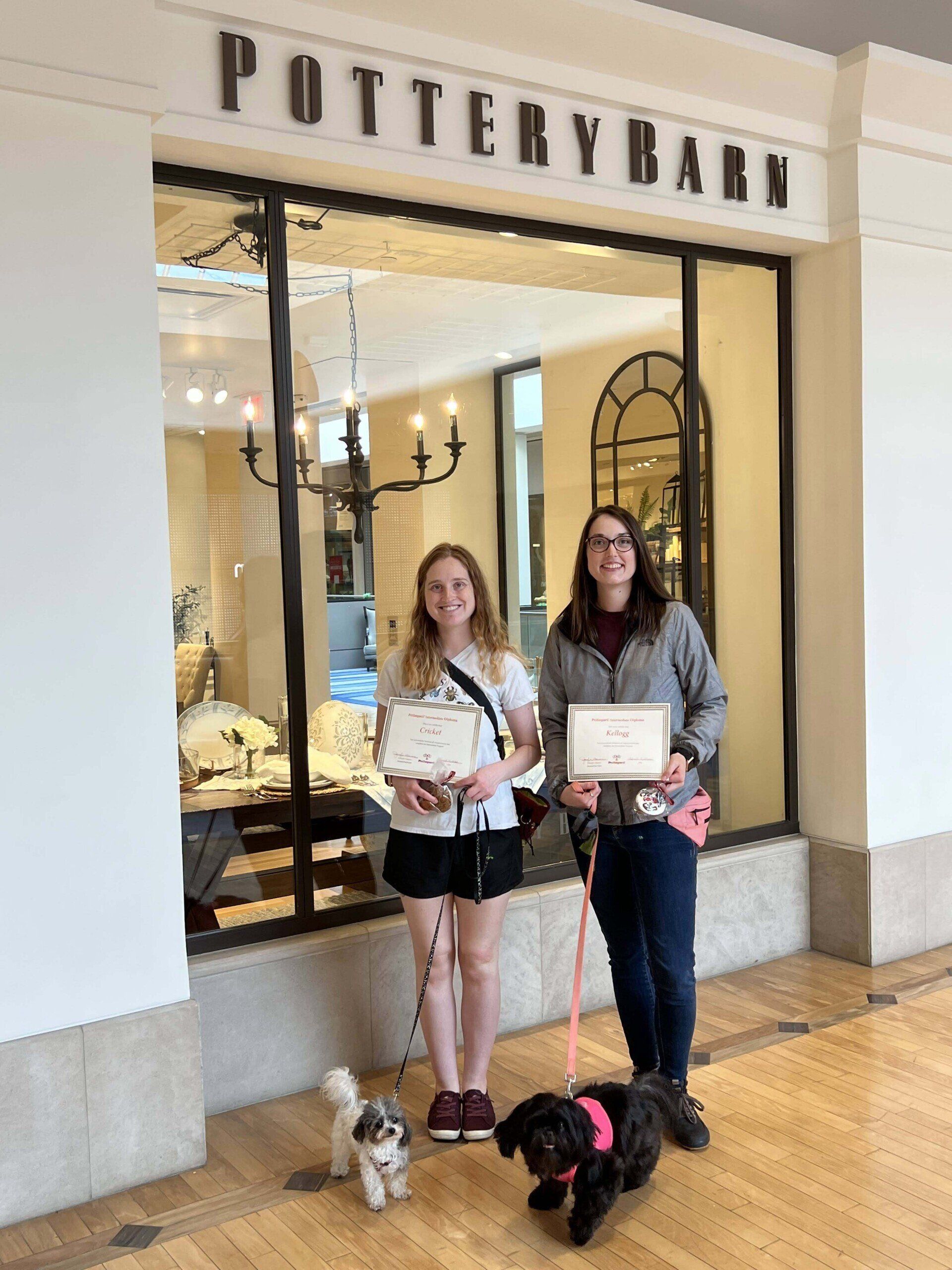 Two people with dogs hold certificates in front of Pottery Barn store.