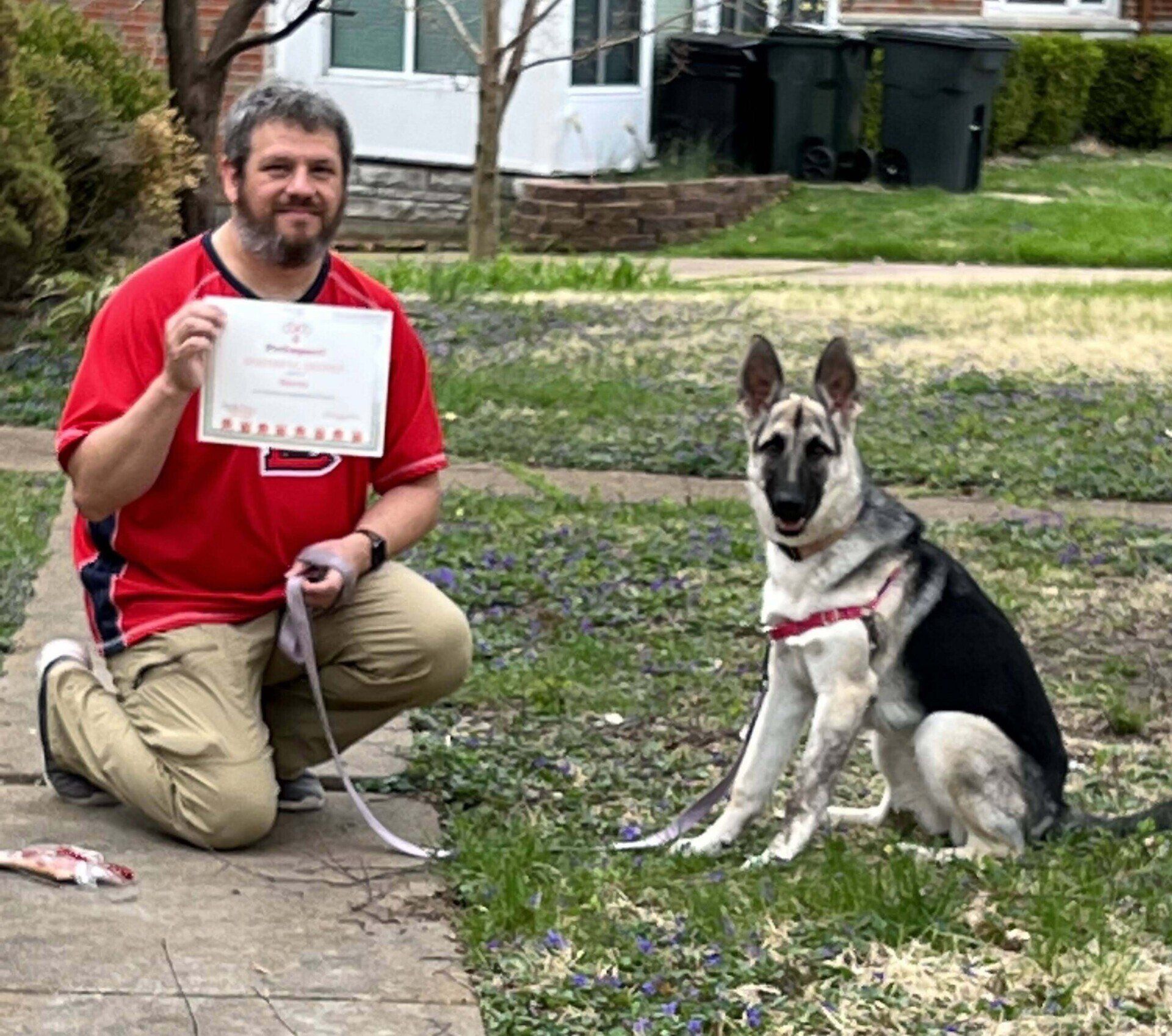 Man kneels next to German Shepherd, holding certificate and leash outside on a path.