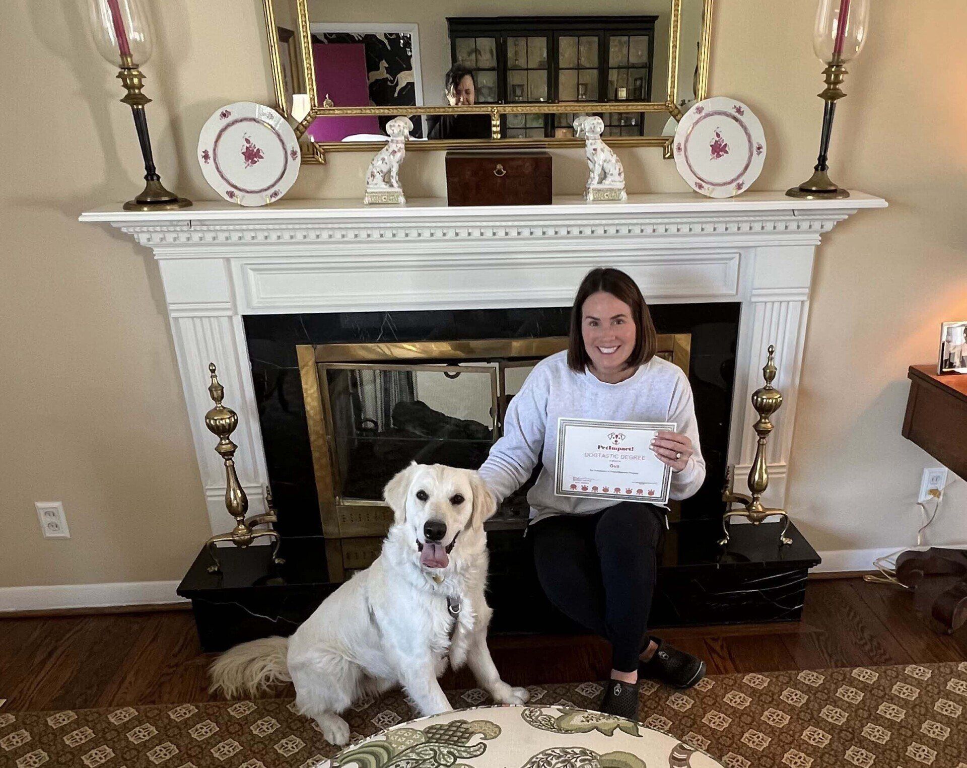 Woman and golden retriever sit in front of fireplace, holding a certificate.