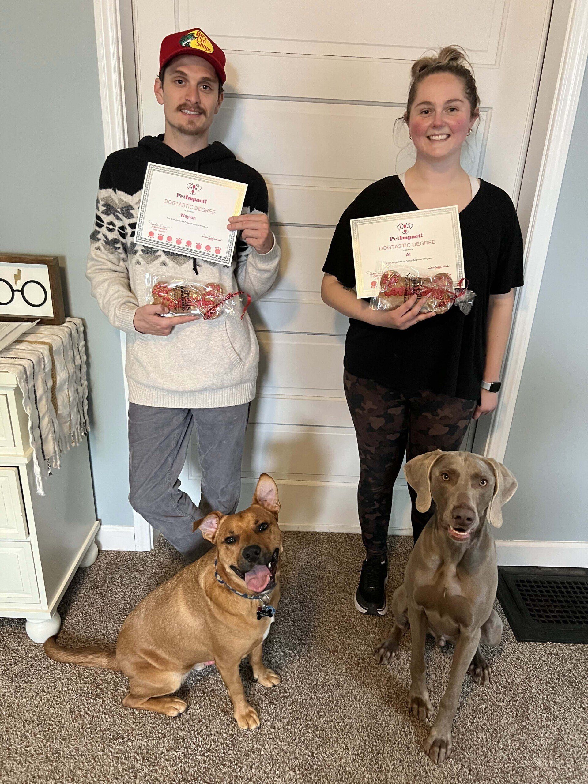 Two people hold certificates with two dogs in a doorway. One person wears a hat.