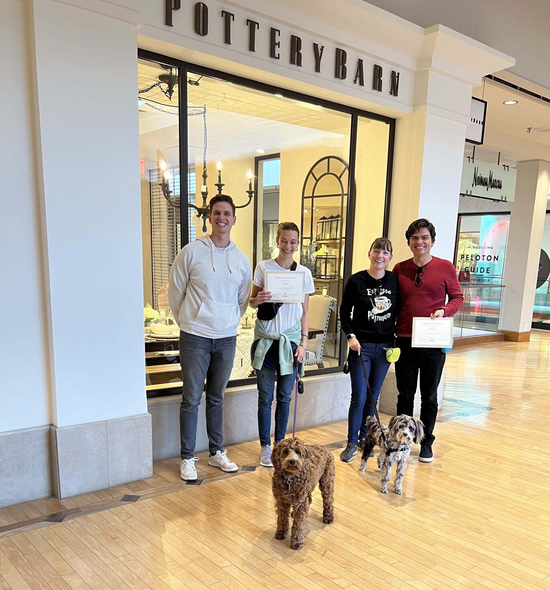 Four people and two dogs pose in front of Pottery Barn in a mall, holding certificates.