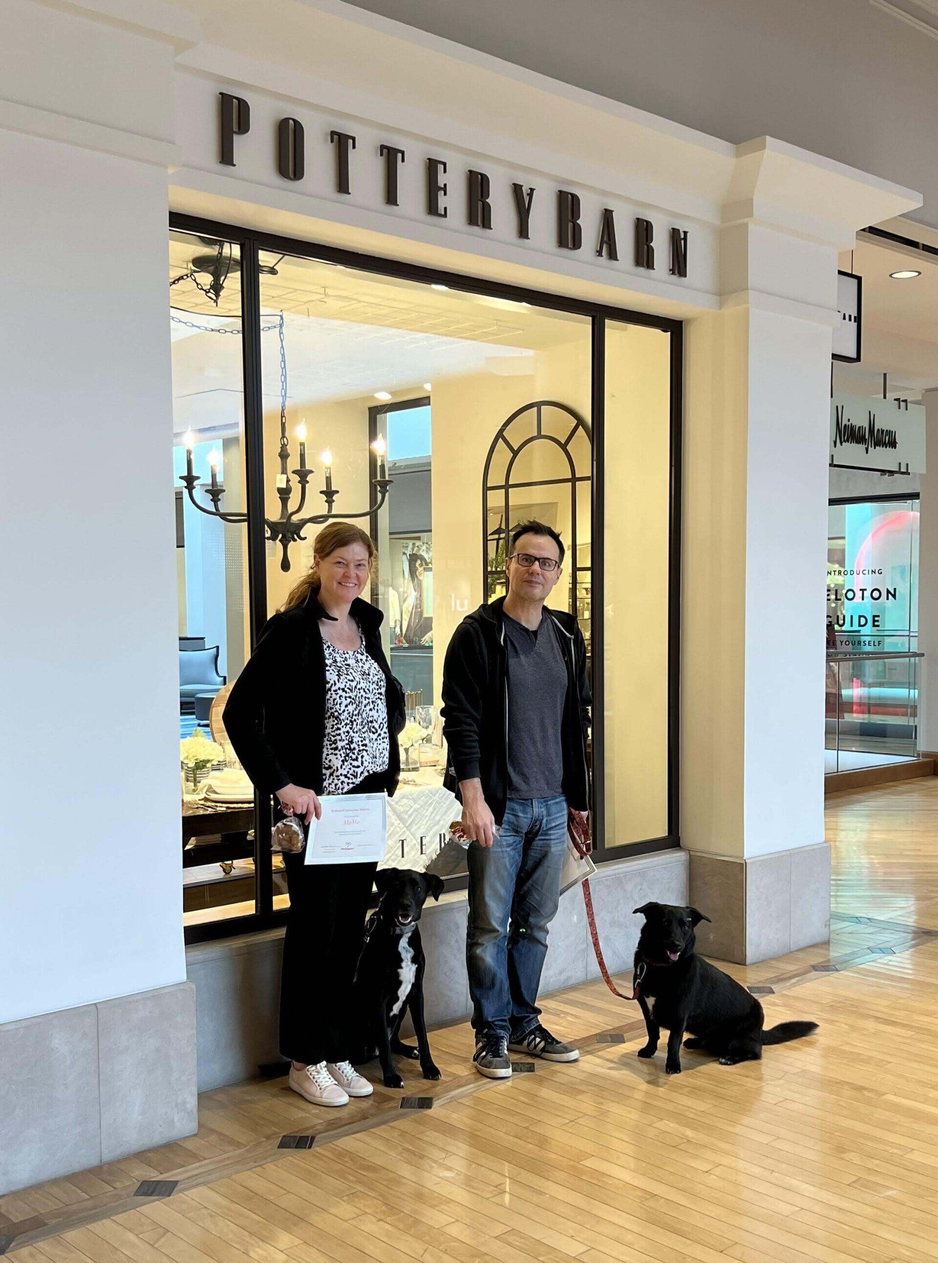 Two people stand outside a Pottery Barn store with two black dogs on leashes.