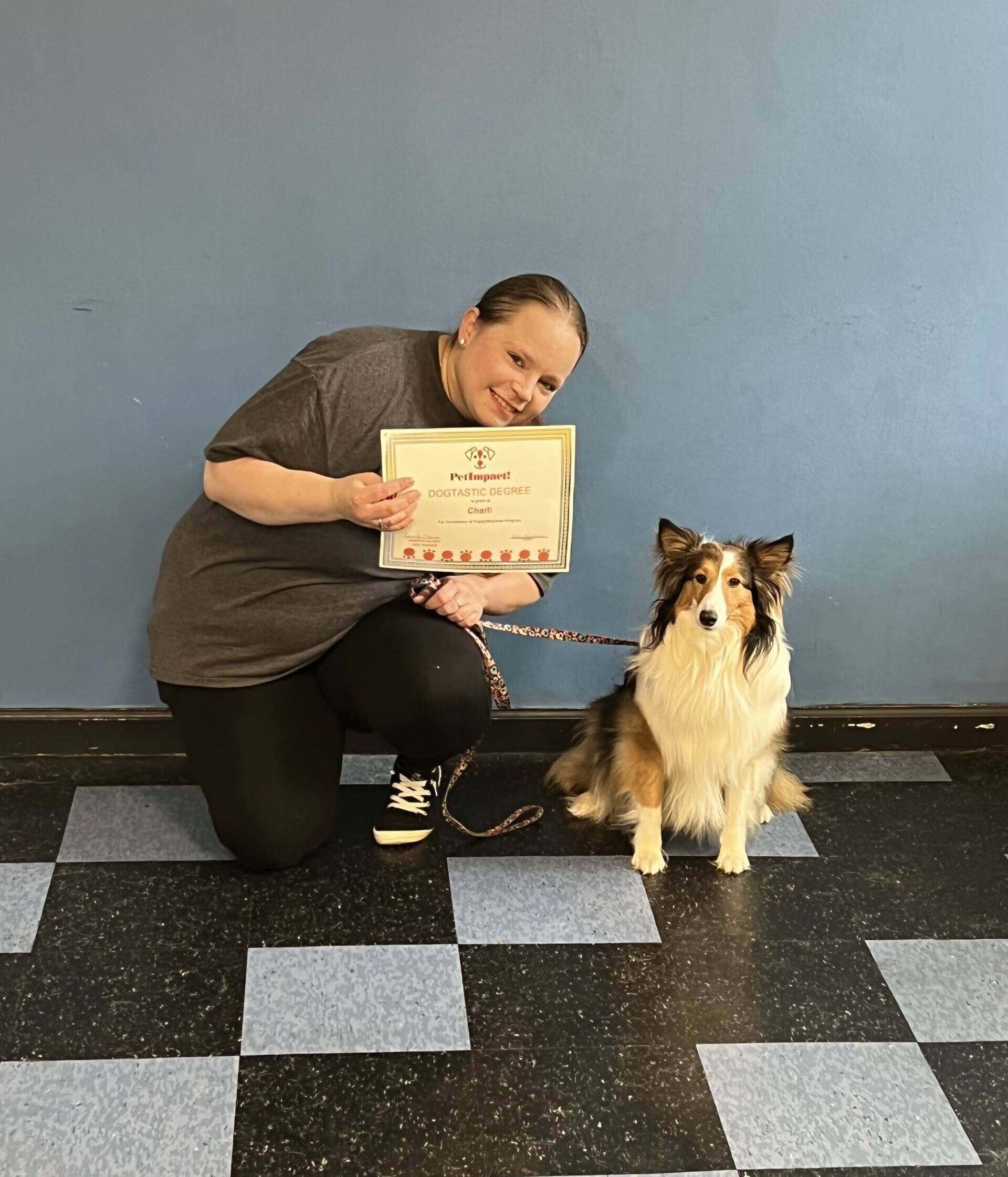 Woman kneeling with dog, holding certificate in front of blue wall. Dog is tri-color, sitting. Smiling.