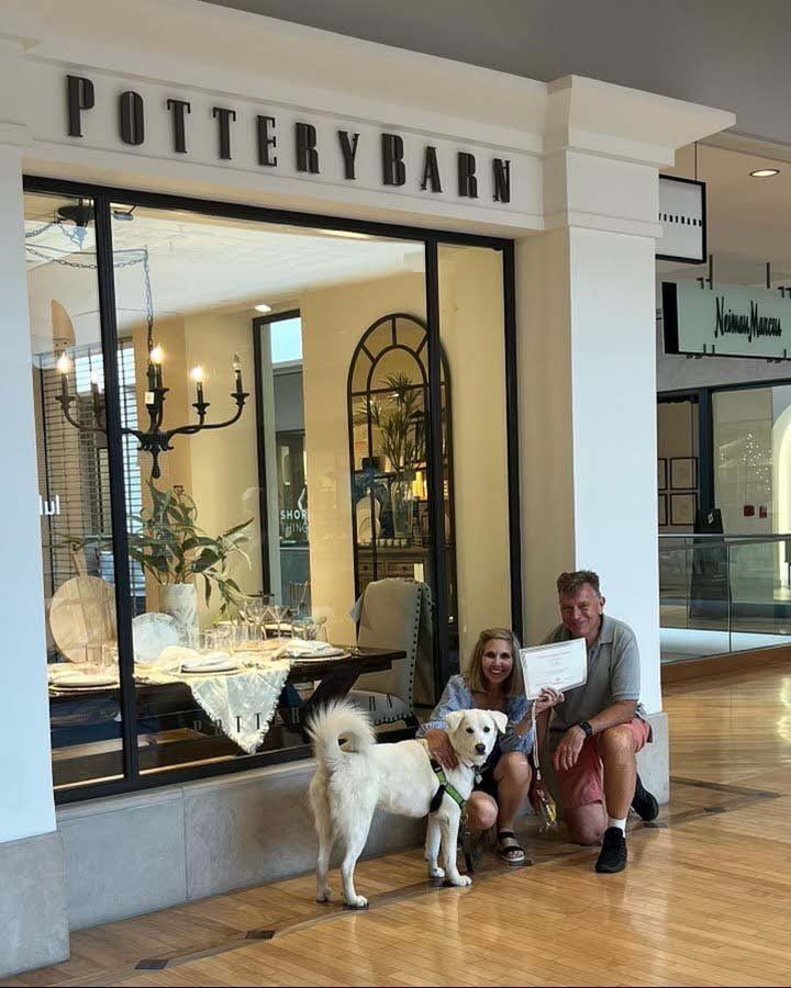 People and dog pose in front of Pottery Barn store. Inside, a table is set.