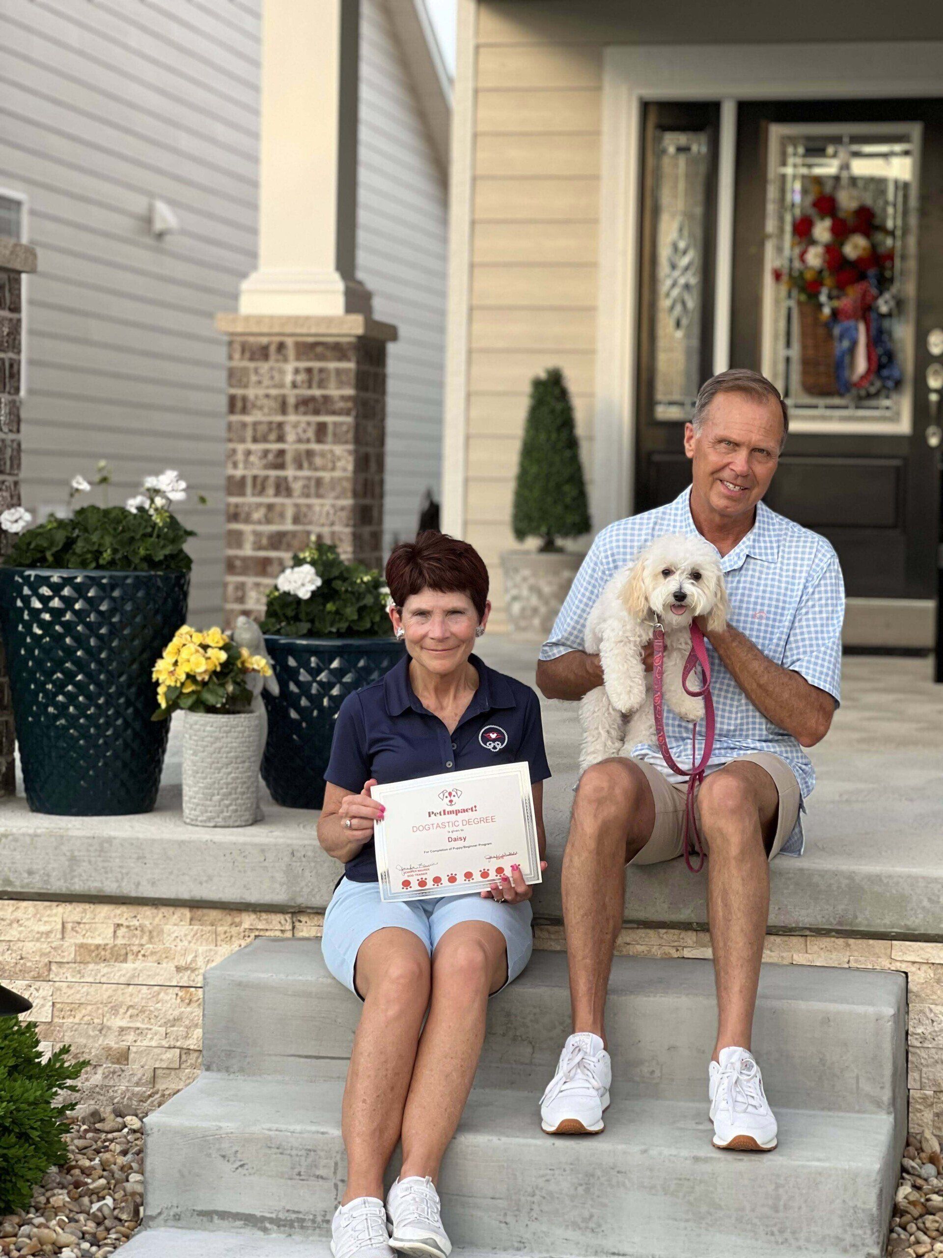 Couple sitting on steps holding a small dog and a sign; front porch with patriotic decorations.