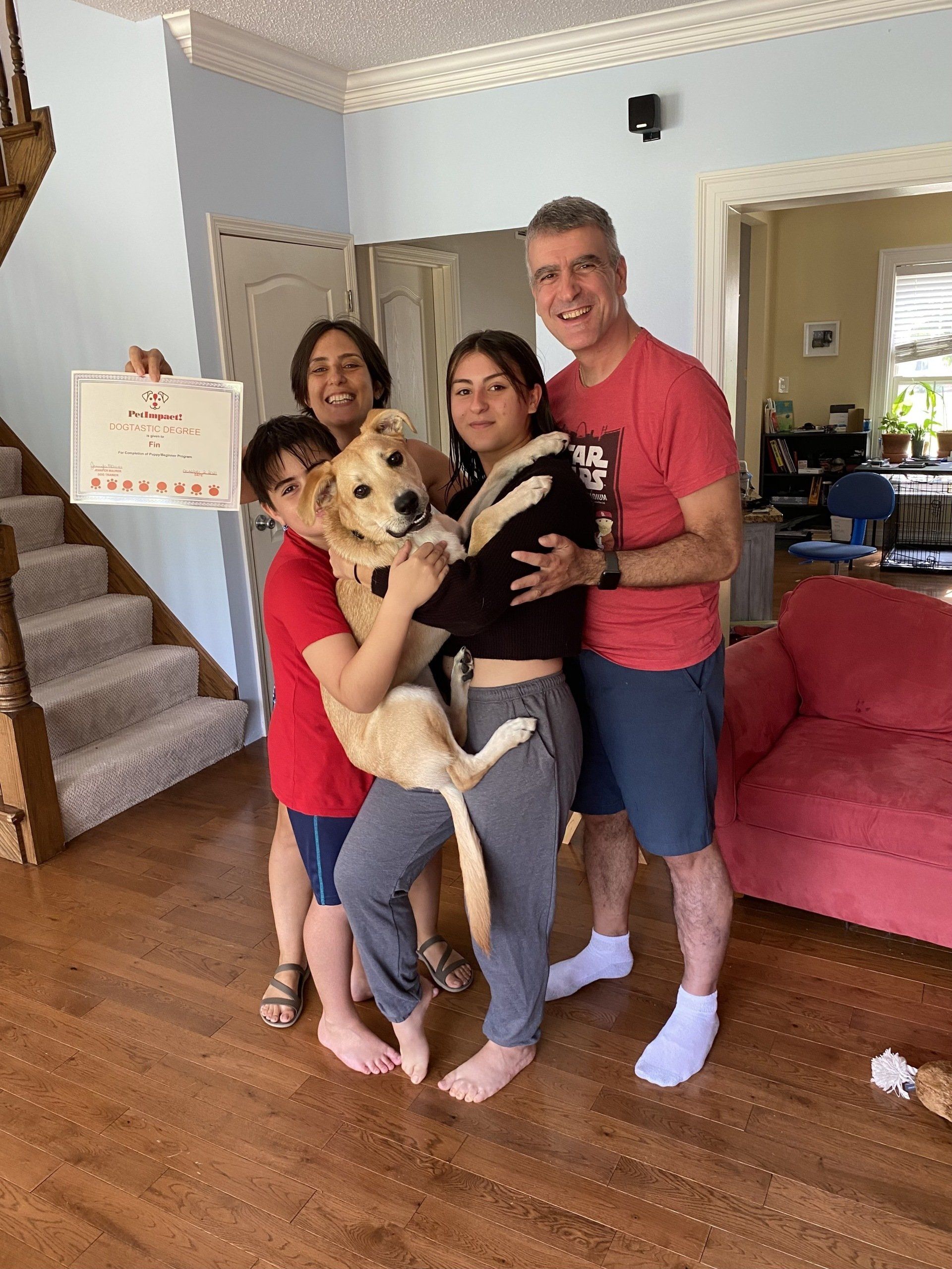 Family of four with a dog, holding a certificate, inside a home, smiling.
