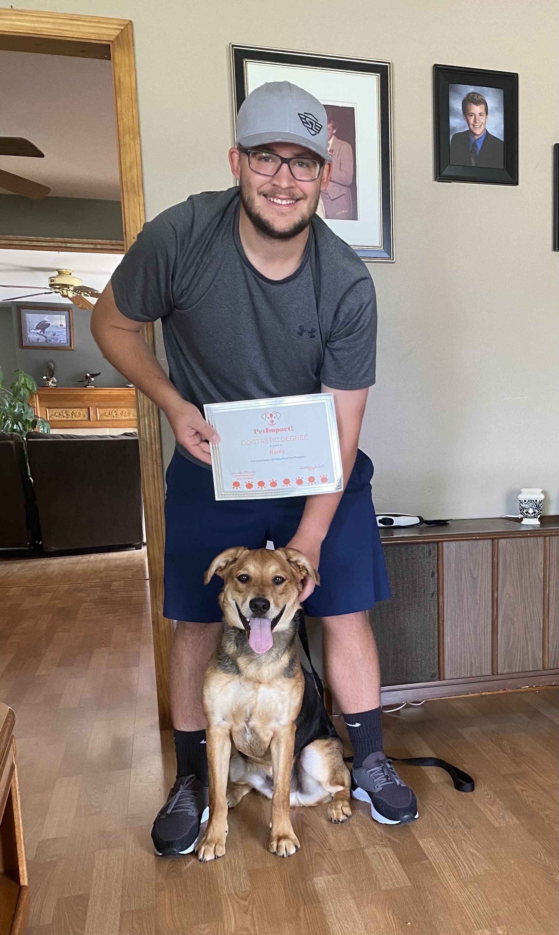 Man and dog pose together, man holding a sign. They're indoors. Dog is brown and tan. Man wearing hat, shorts, and gray shirt, smiling.