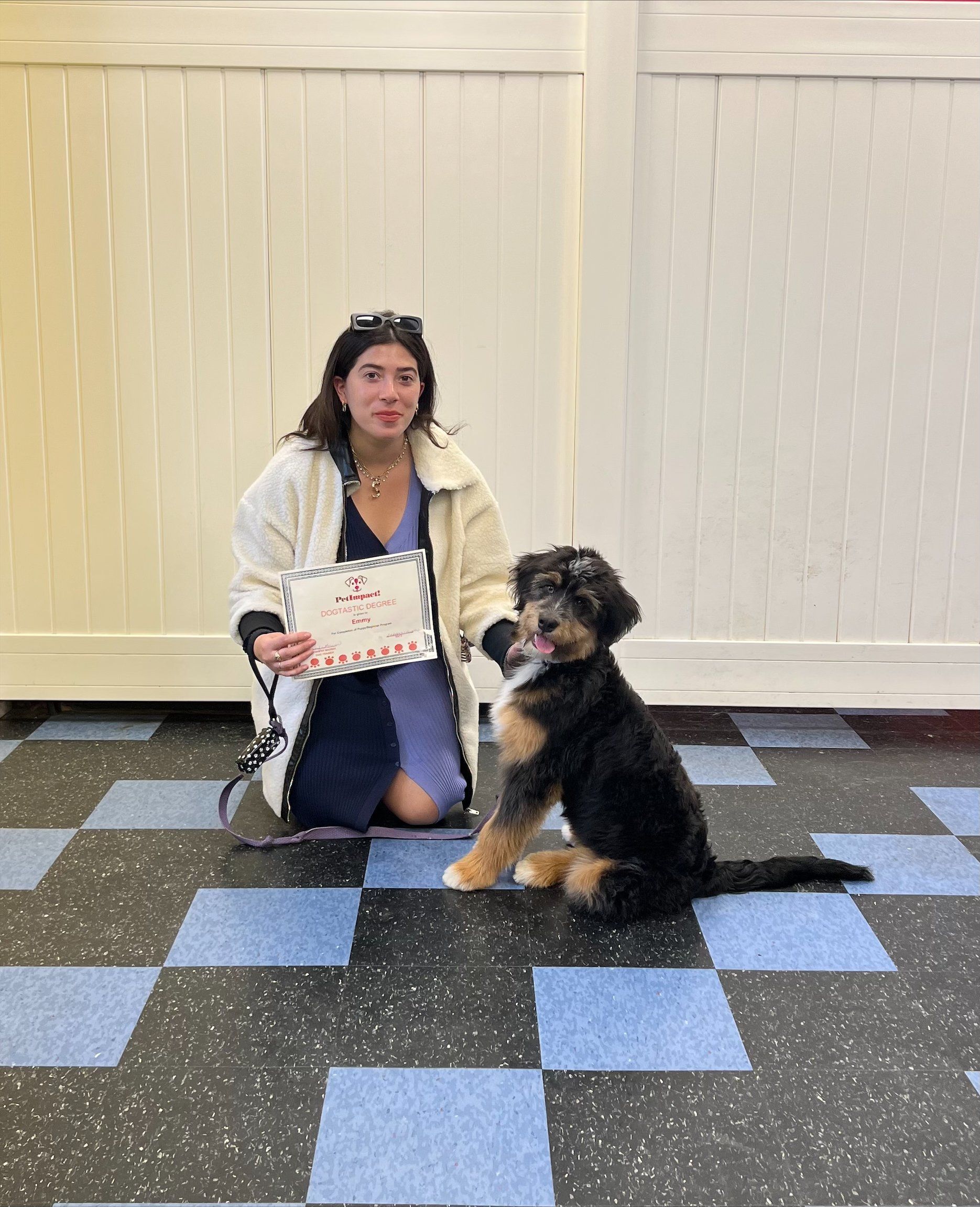Woman kneels next to a black and brown dog, both on a checkered floor, holding a sign.