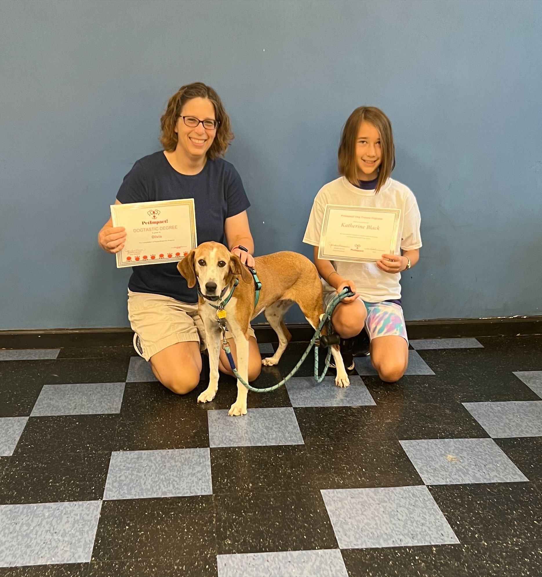 Woman and girl with a dog, holding certificates, kneeling on checkered floor. Blue wall backdrop.