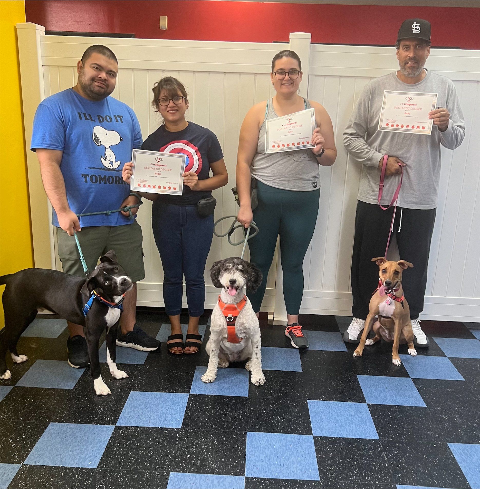 Four people with leashed dogs hold certificates, posing in a dog training area with blue and black tiled floor and a white fence.