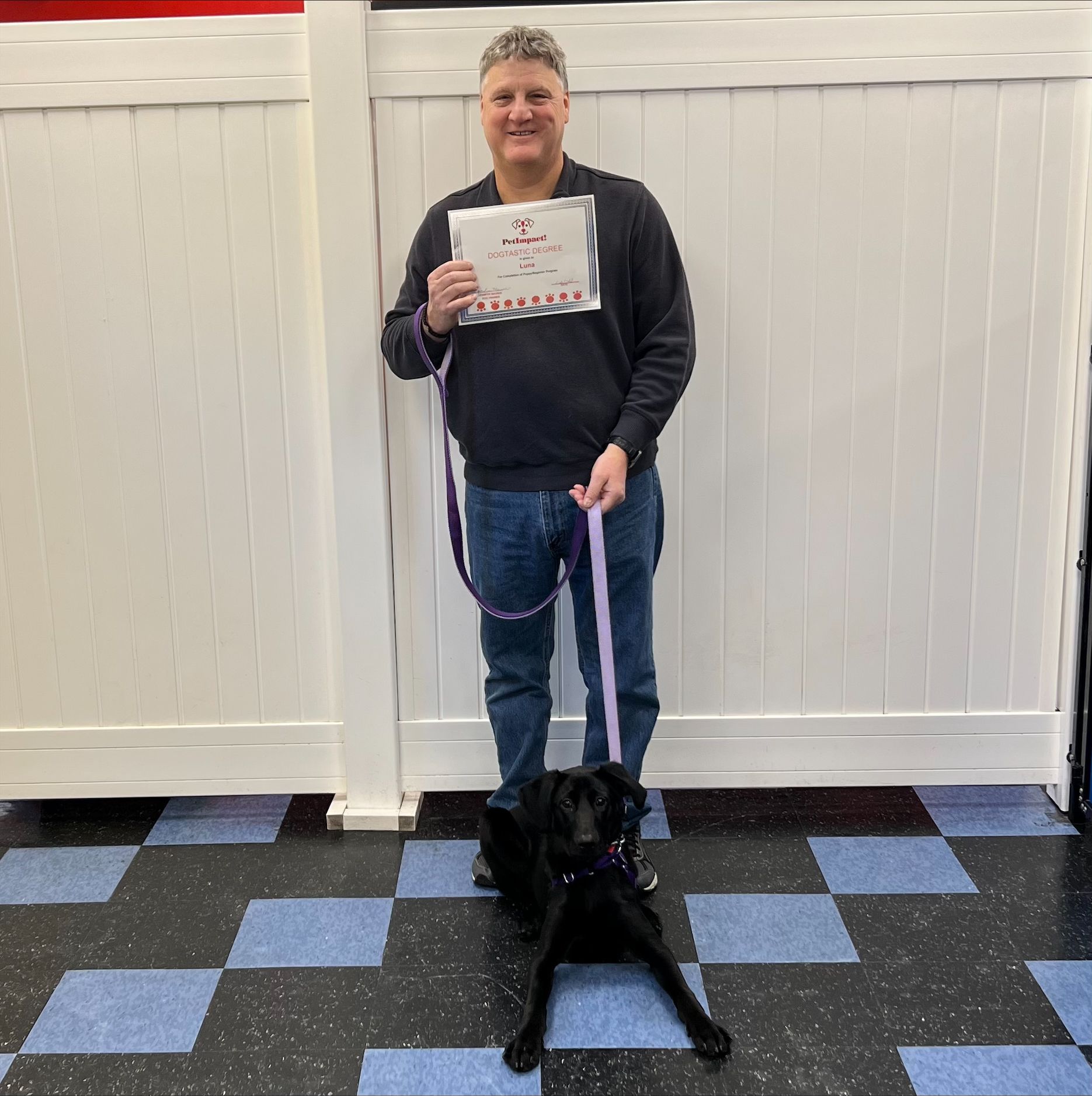 Man and black dog pose, holding certificate, dog on leash, in a room with patterned floor.