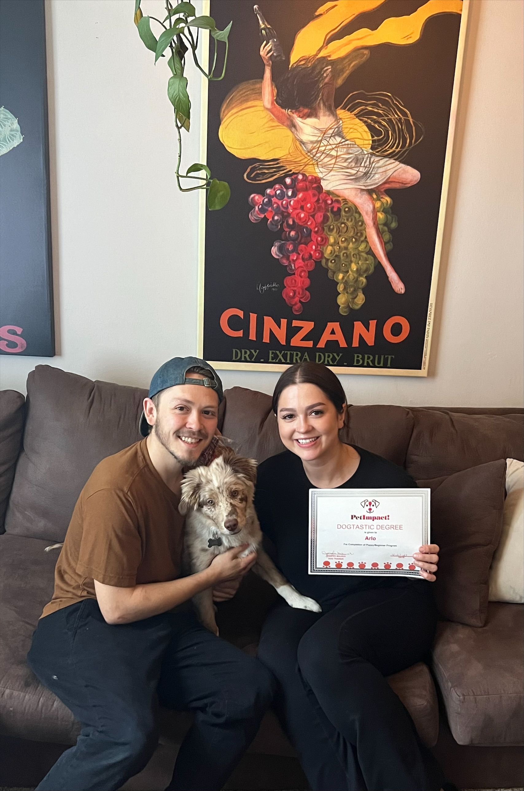 Couple and dog sit on a brown couch holding a framed certificate. A Cinzano poster hangs behind them.