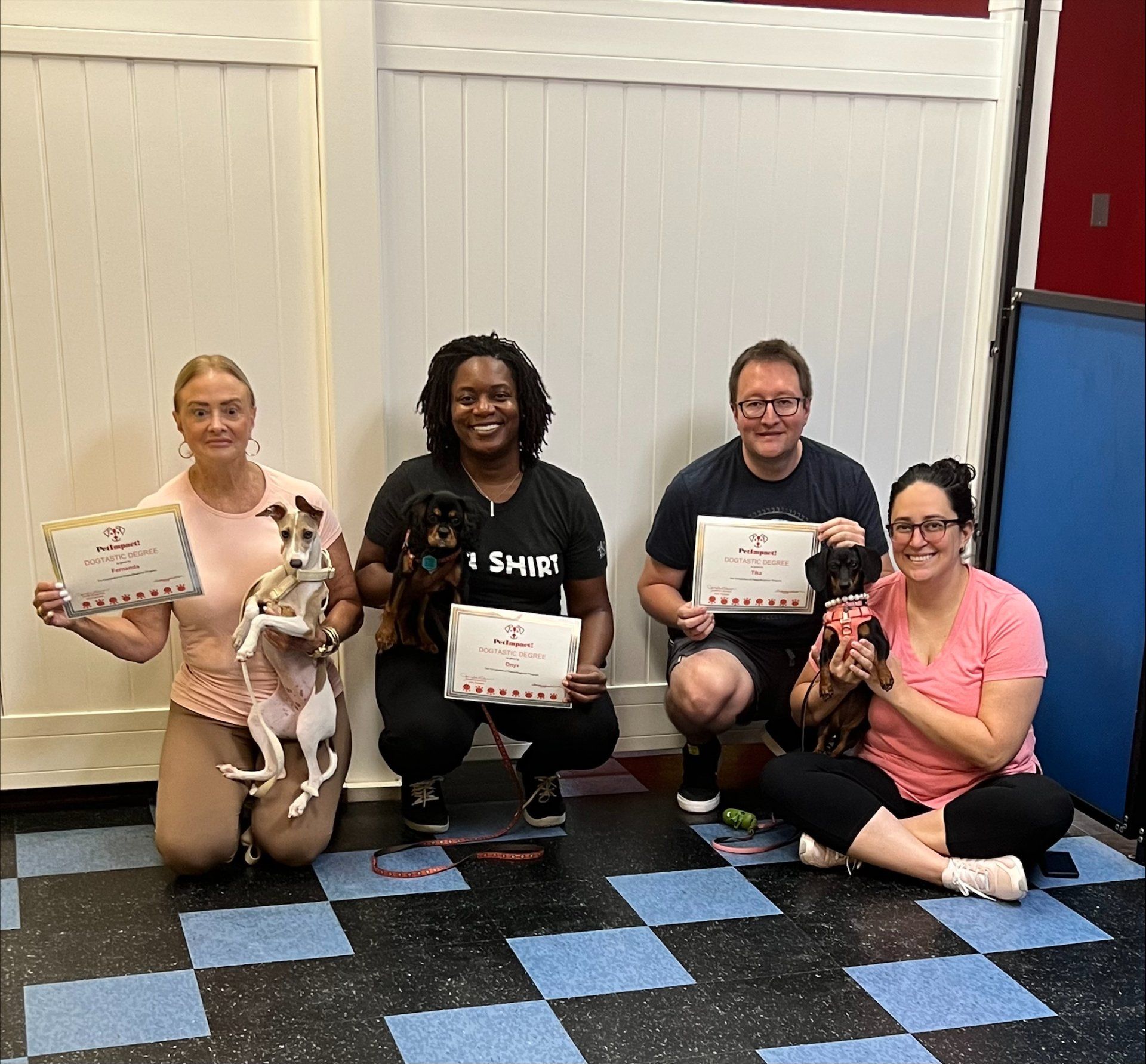 Four people with dogs hold certificates, posing in front of a white wall.