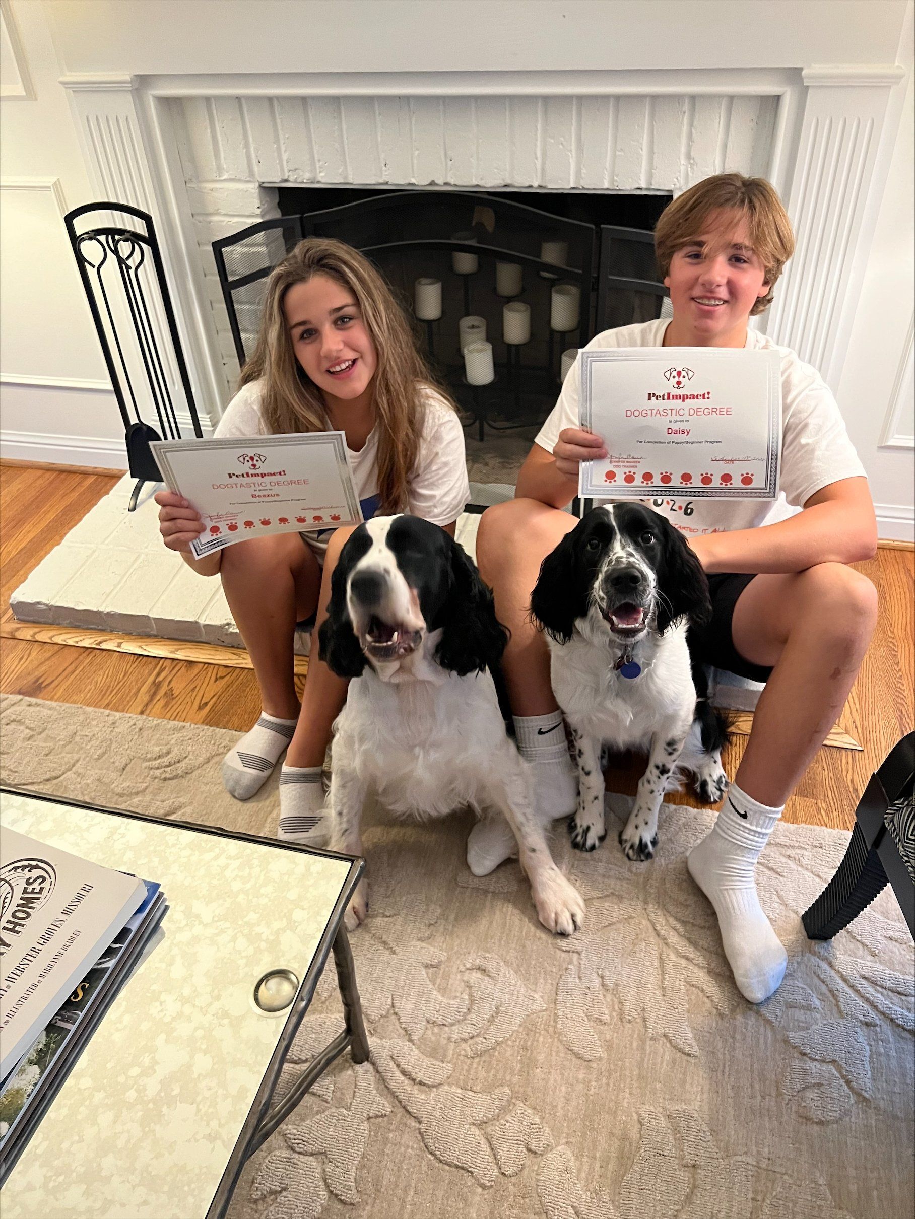 Two people and two dogs holding certificates by a fireplace; all smiling.