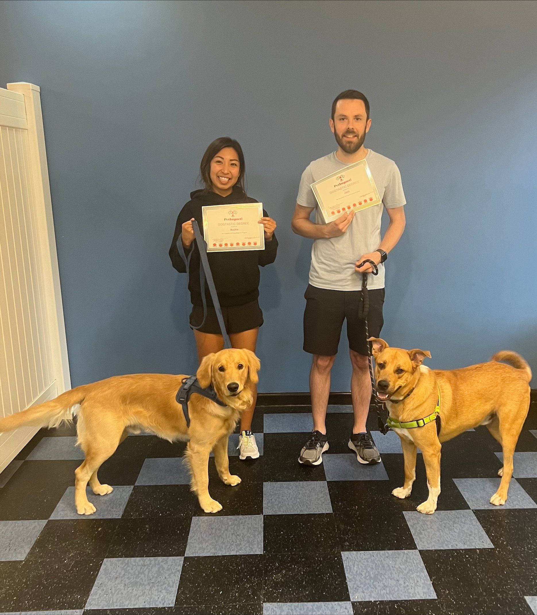 Two people and two dogs pose with certificates in front of a blue wall. Dogs are on leashes.
