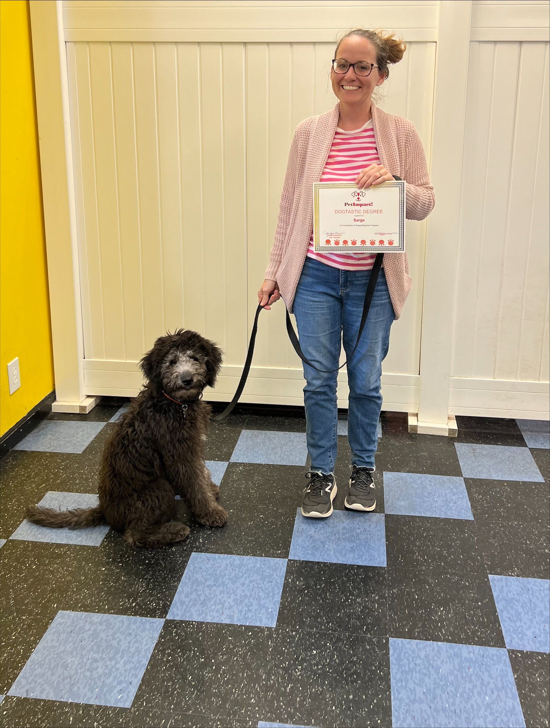 Woman with dog holds certificate on checkered floor, in front of a white wall.