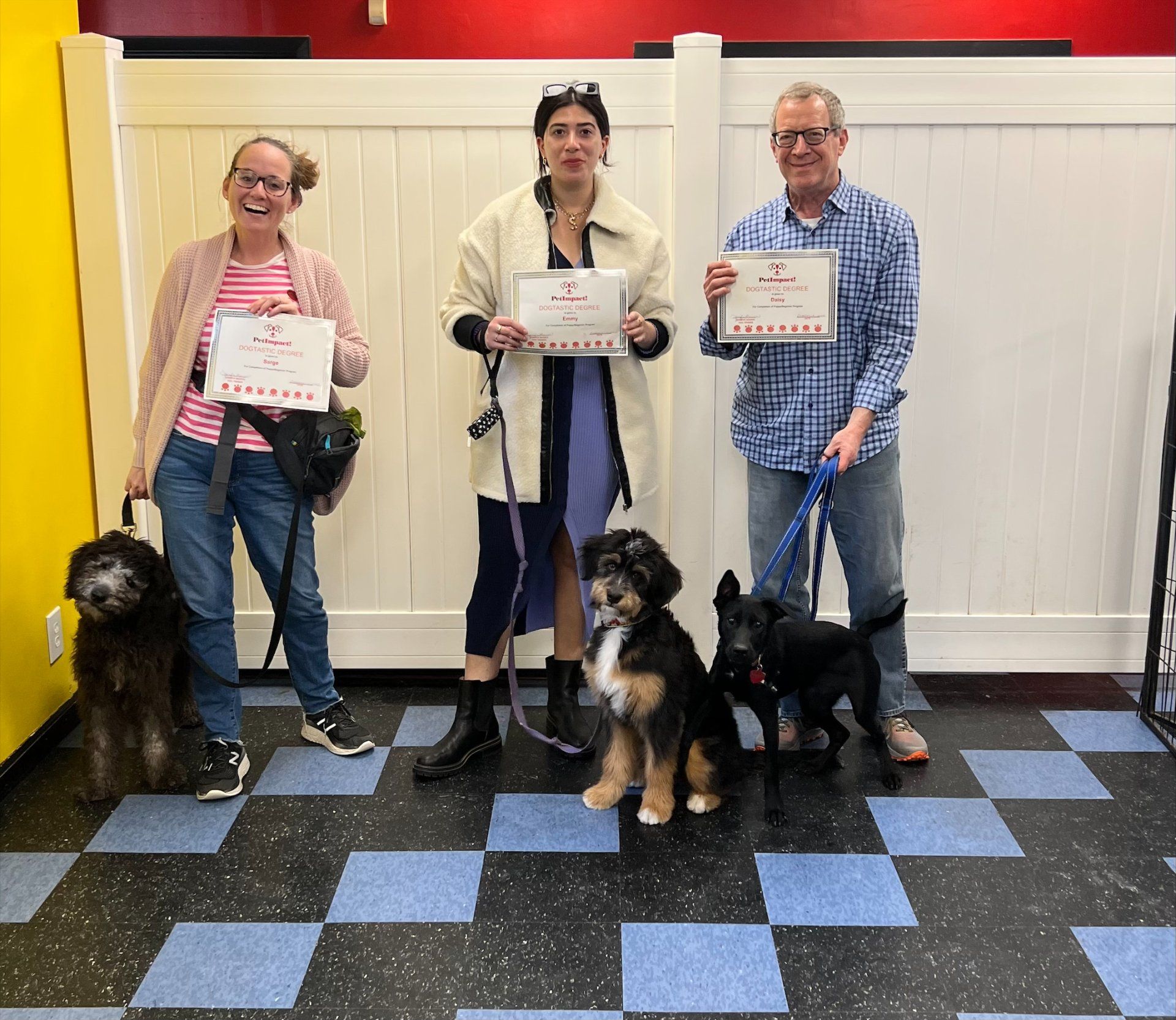 Three people with dogs pose with certificates. They stand in a dog training area with checkered floors.