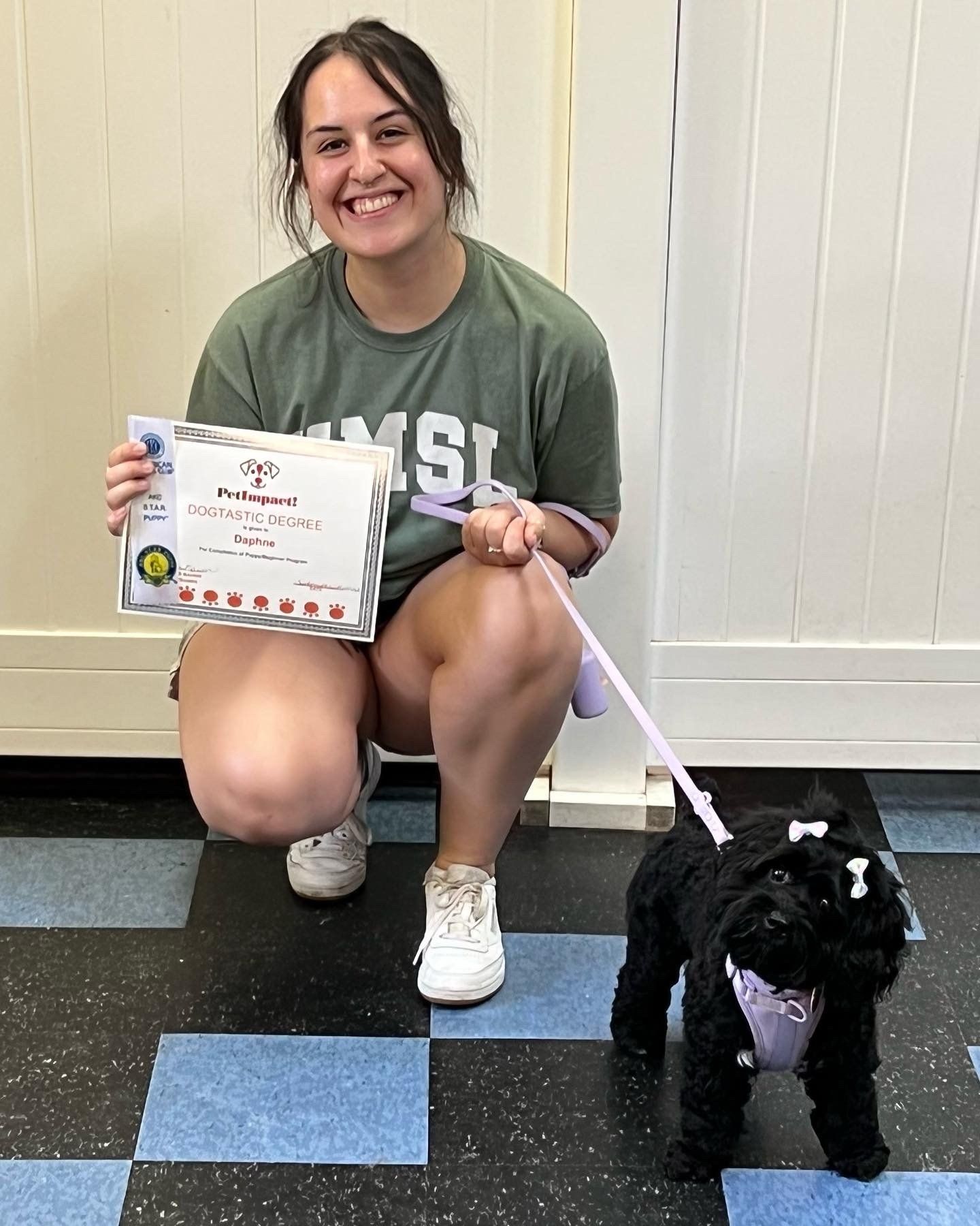 Woman kneels with a small black dog on a leash, holding a certificate.