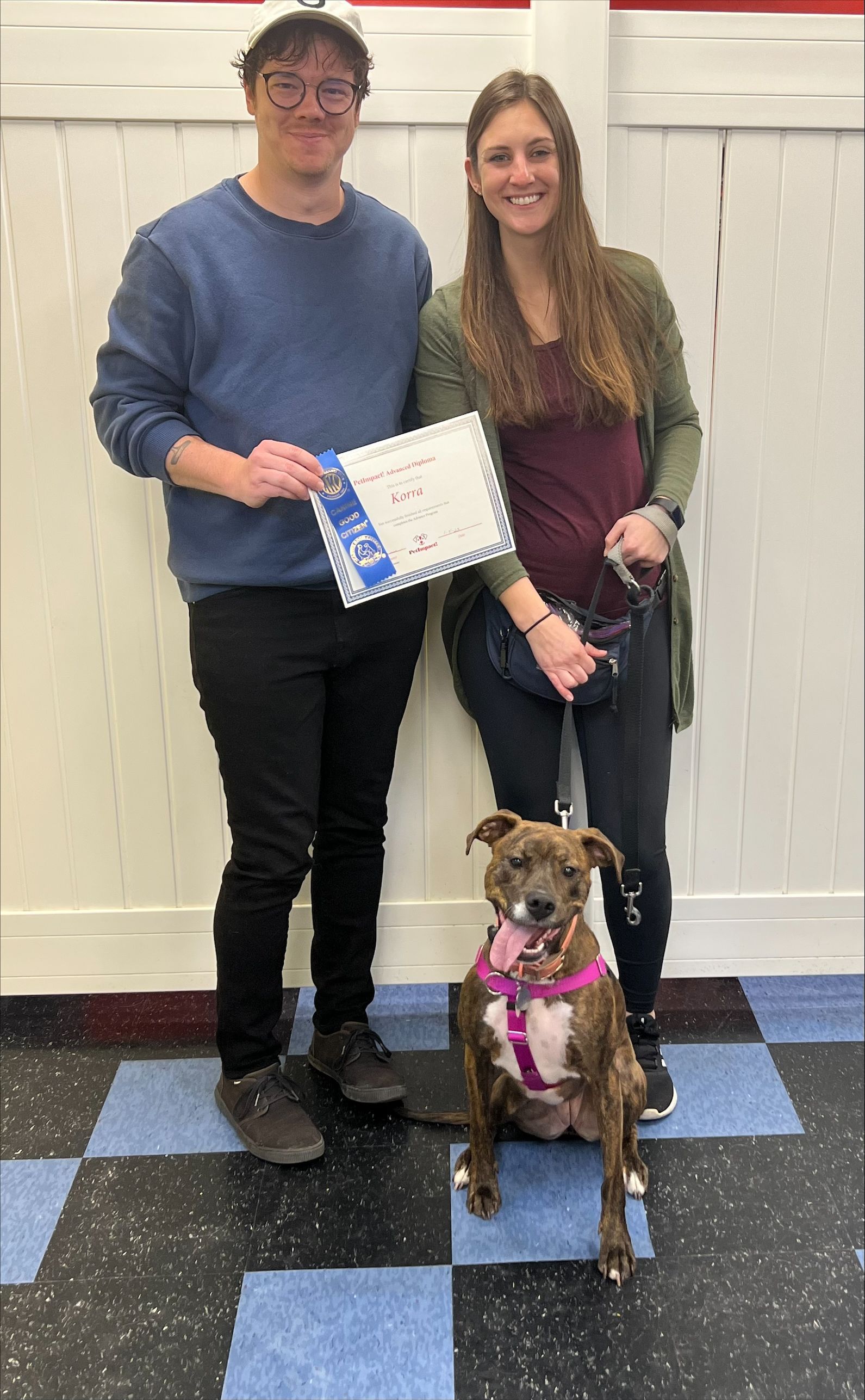 Couple with a brown dog, holding a certificate. The dog wears a pink harness, indoors with a checkered floor.