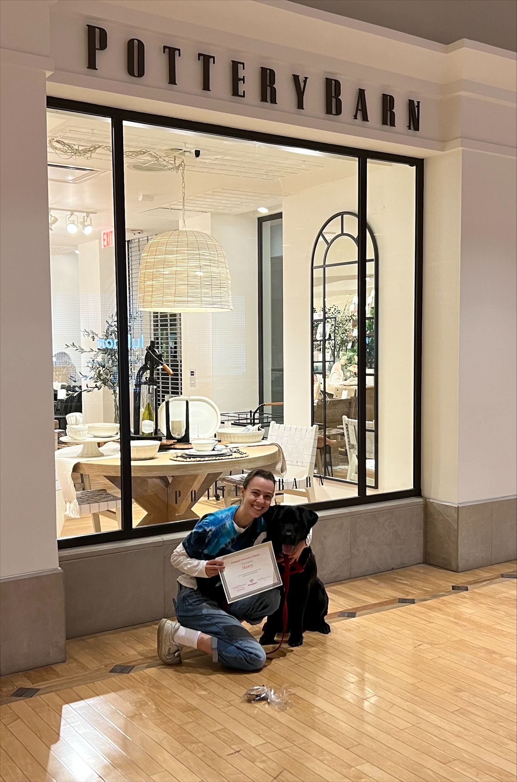 Woman kneeling with black dog in front of Pottery Barn store. Smiling, holding a sign.
