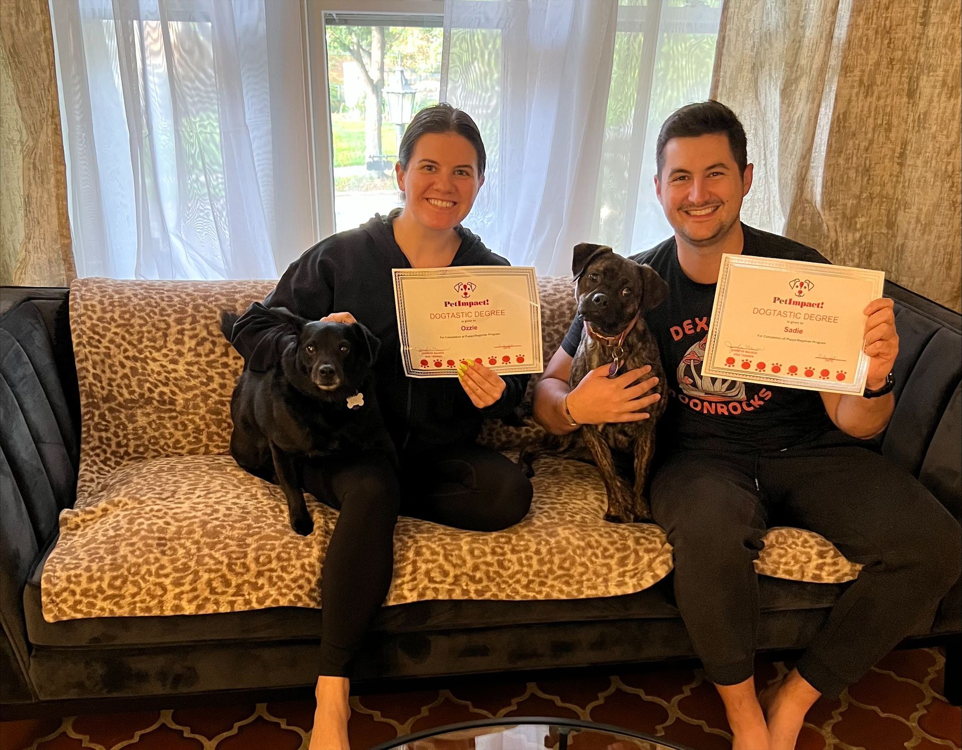 Two people on a couch with two dogs, holding awards. Smiling, indoors.