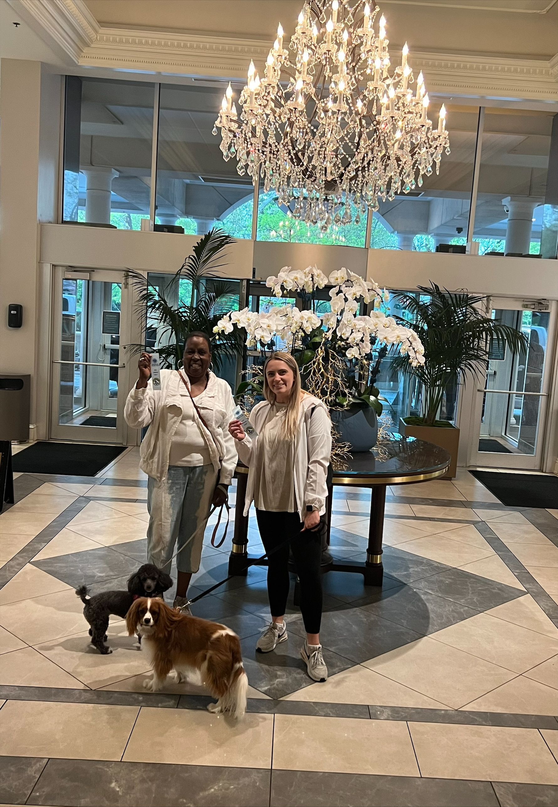 Two women and two dogs in a hotel lobby with a chandelier and floral arrangement.