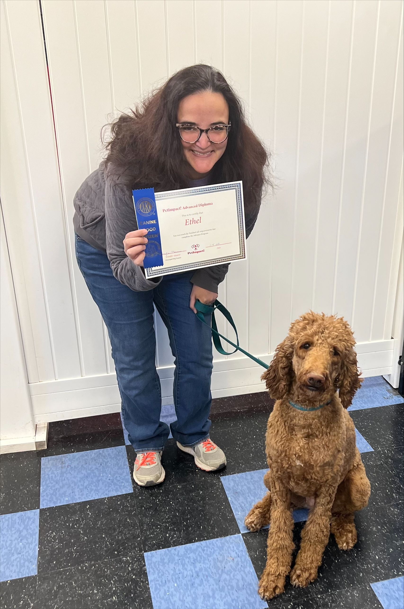 Woman with curly hair and dog smiling, holding a certificate with a blue ribbon.