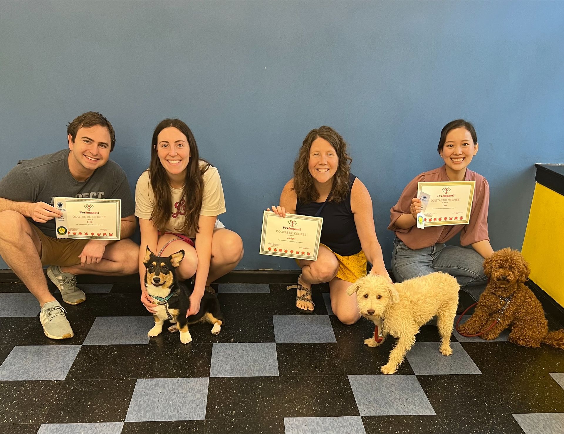 Four people kneel with their dogs, holding certificates in front of a blue wall.
