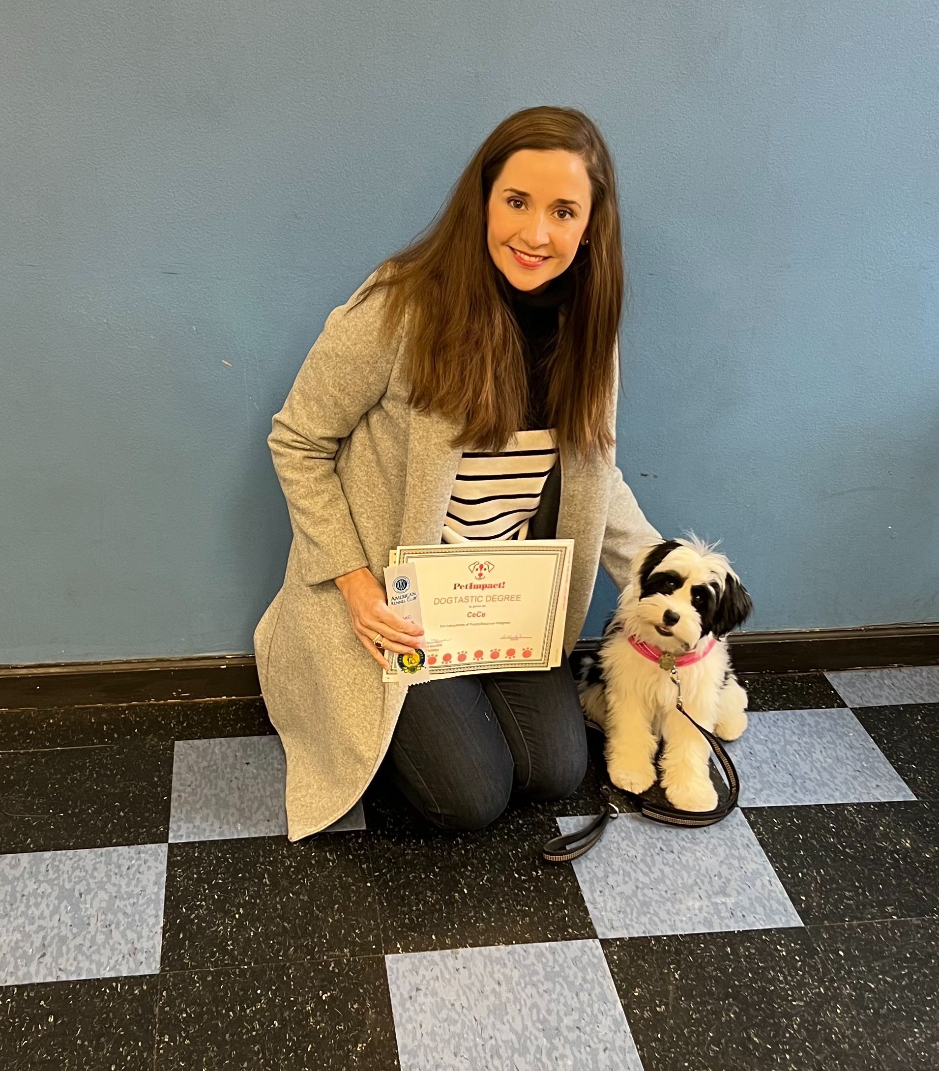 Woman kneeling with a small dog, holding a certificate. Blue wall and checkered floor in the background.