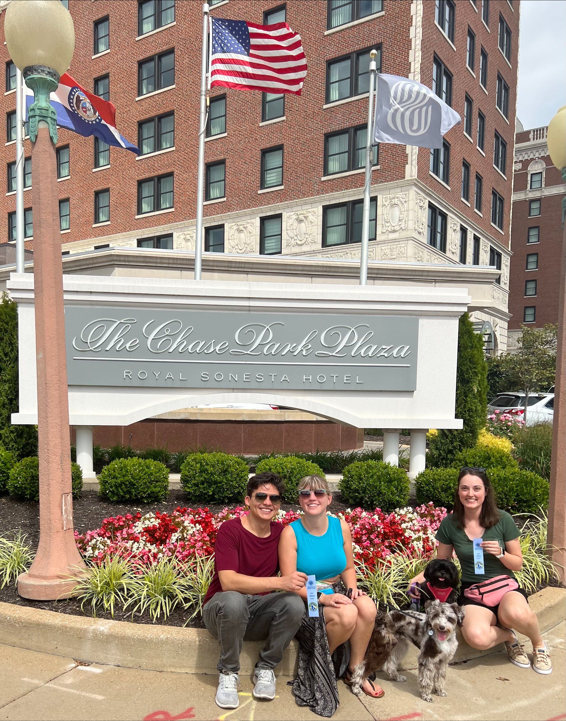 Three women and two dogs in front of The Chase Park Plaza sign; U.S., Missouri flags in background.