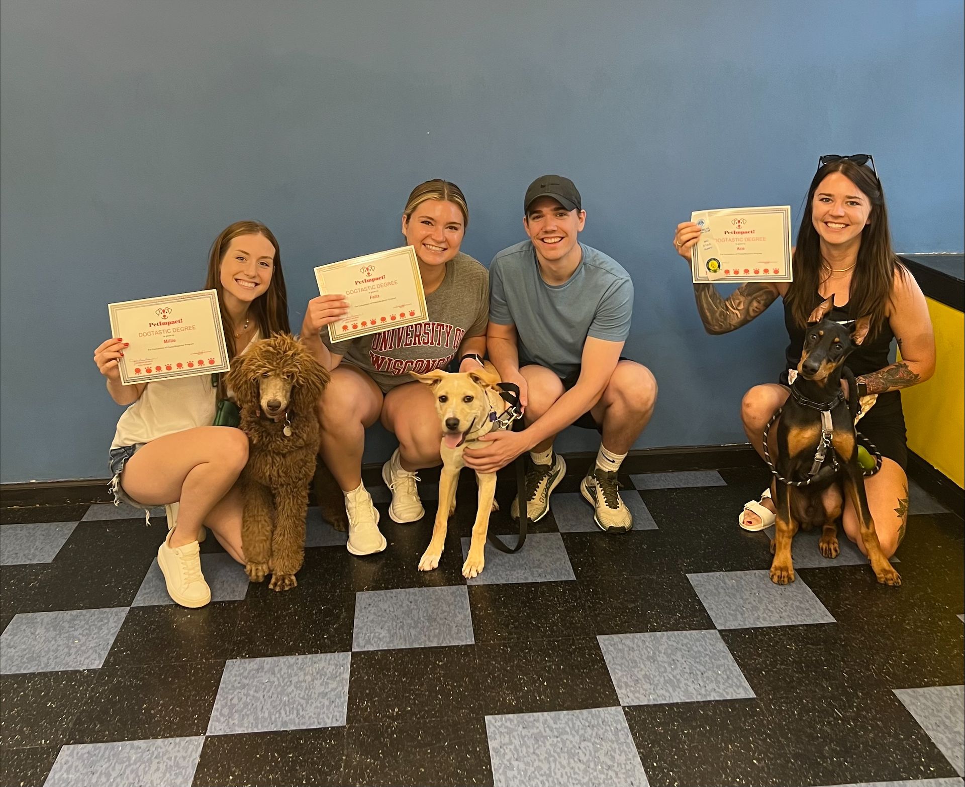 People with dogs holding certificates in front of a blue wall and checkered floor.