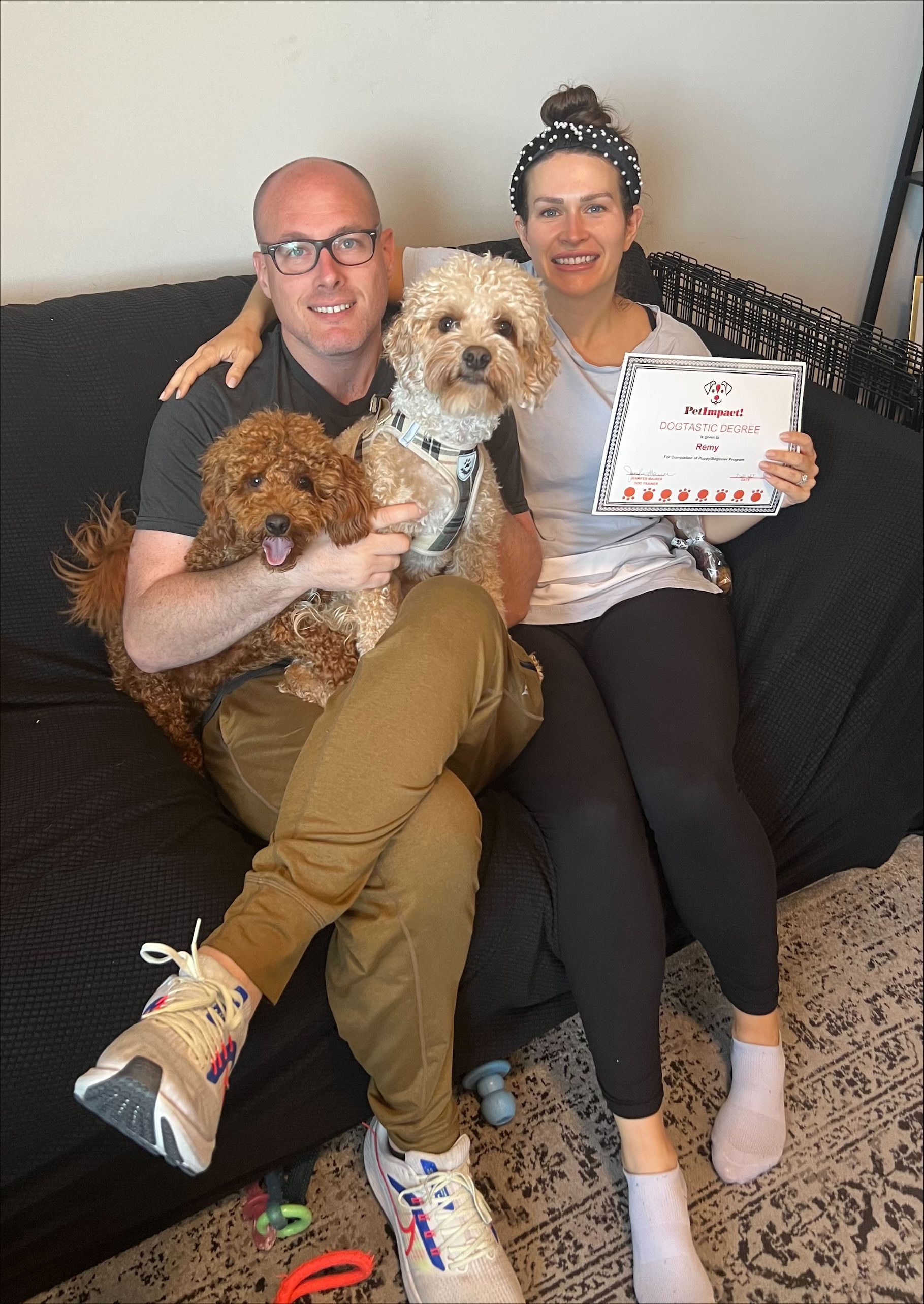 A couple sits on a black couch with two dogs, holding a framed certificate.